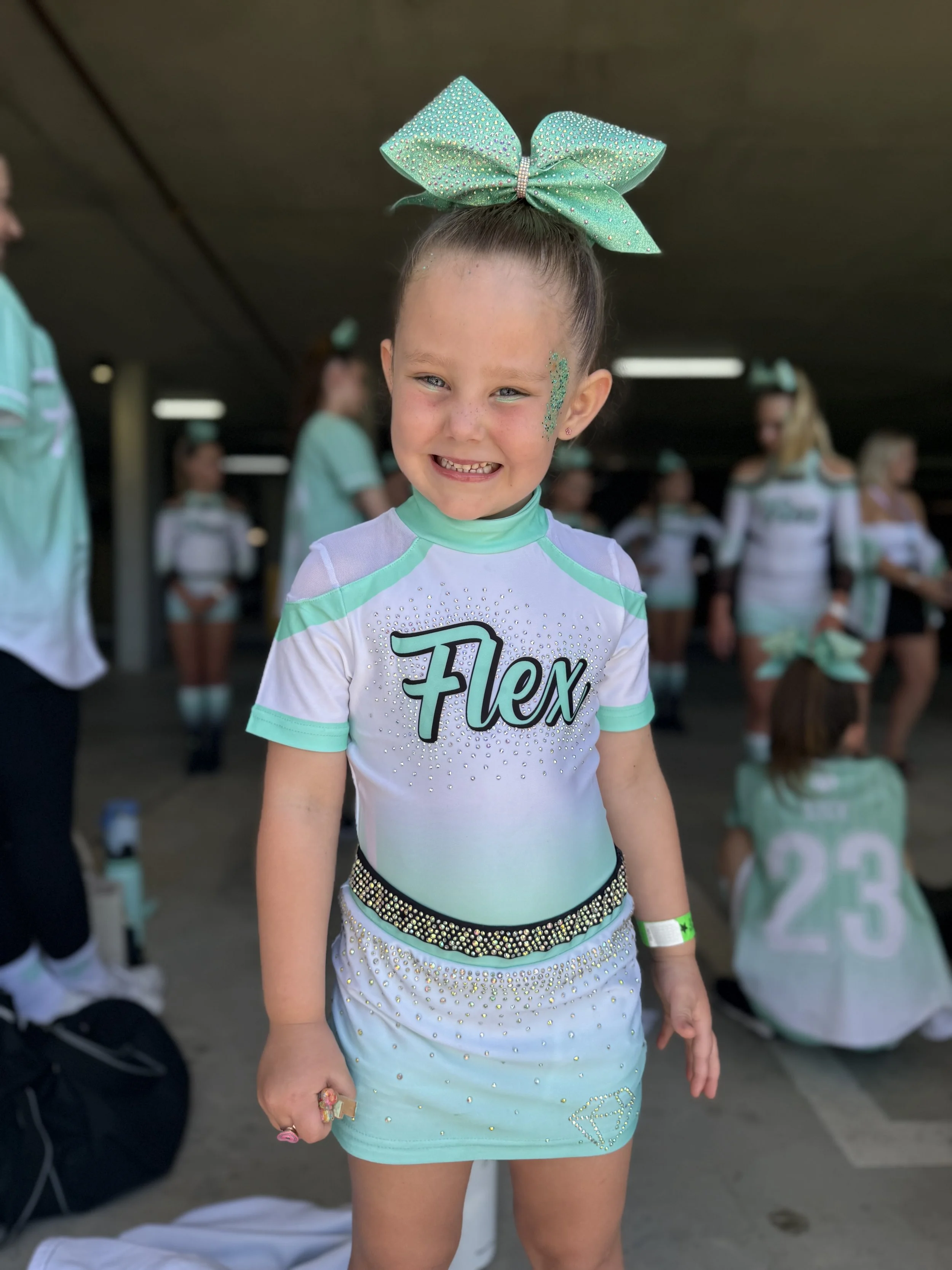 Young girl in a cheerleading uniform with the word 'Flex' on it, smiling with a big green bow in her hair, and glitter on her face, standing in a group of cheerleaders in similar uniforms.