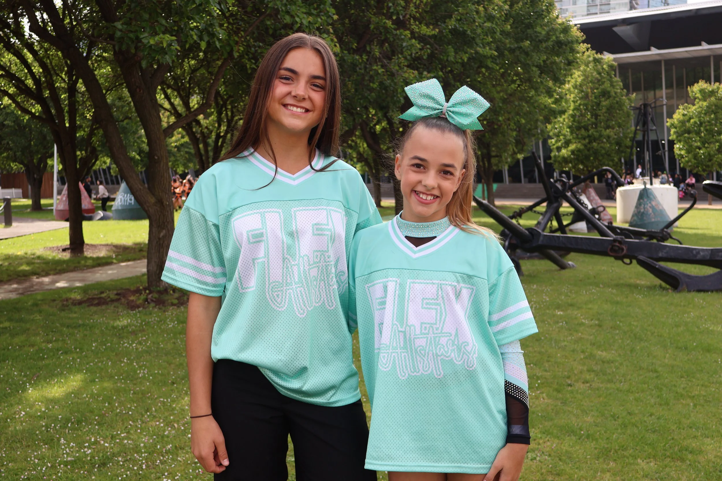 Two young girls standing outdoors in a park, smiling at the camera, wearing matching teal sports jerseys with 'FLEX' written on them, surrounded by green trees and grass.