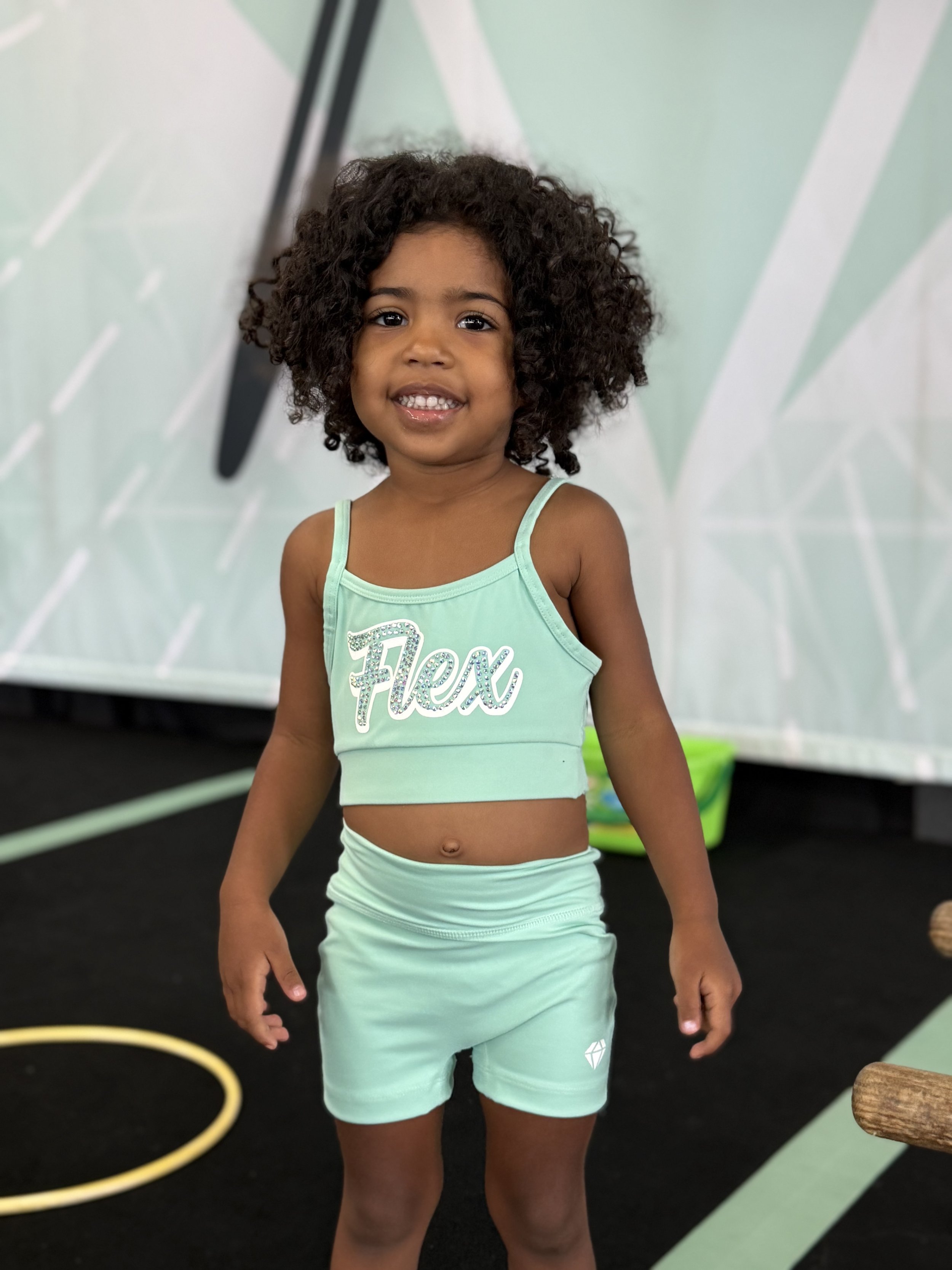 A young girl with curly hair smiling at the camera in a mint green sports outfit with