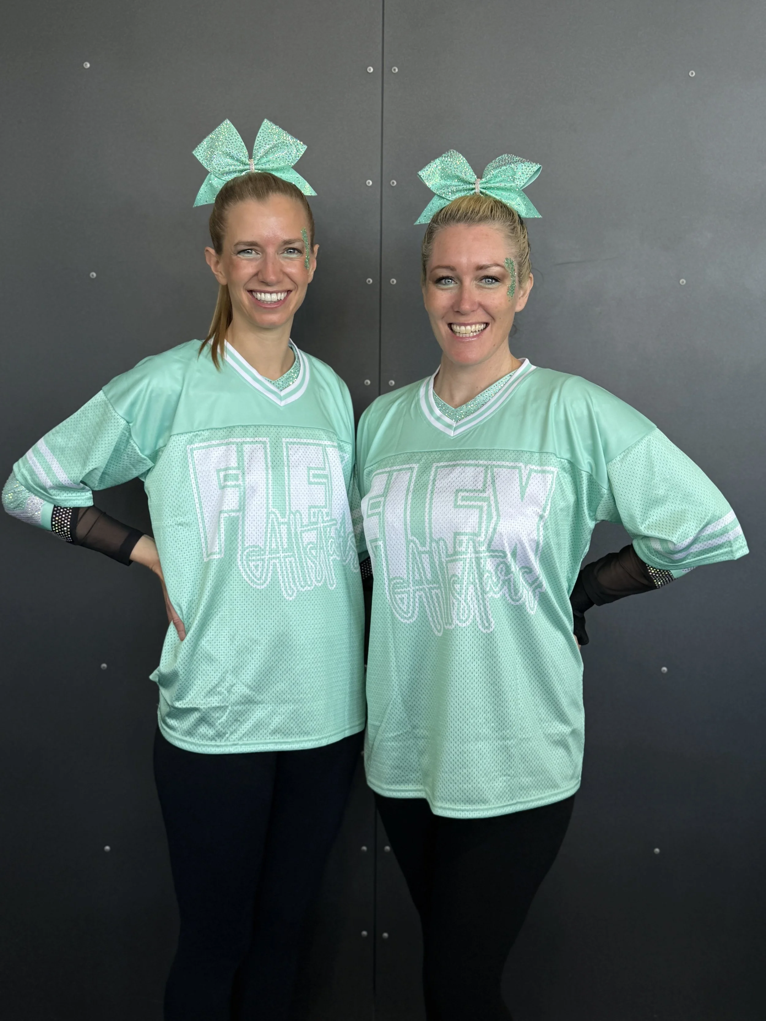 Two women wearing matching turquoise cheerleading jerseys with 'FLEX CHEER' written on them, standing in front of a dark wall with small metal fasteners, both with turquoise bows on their hair and smiling.
