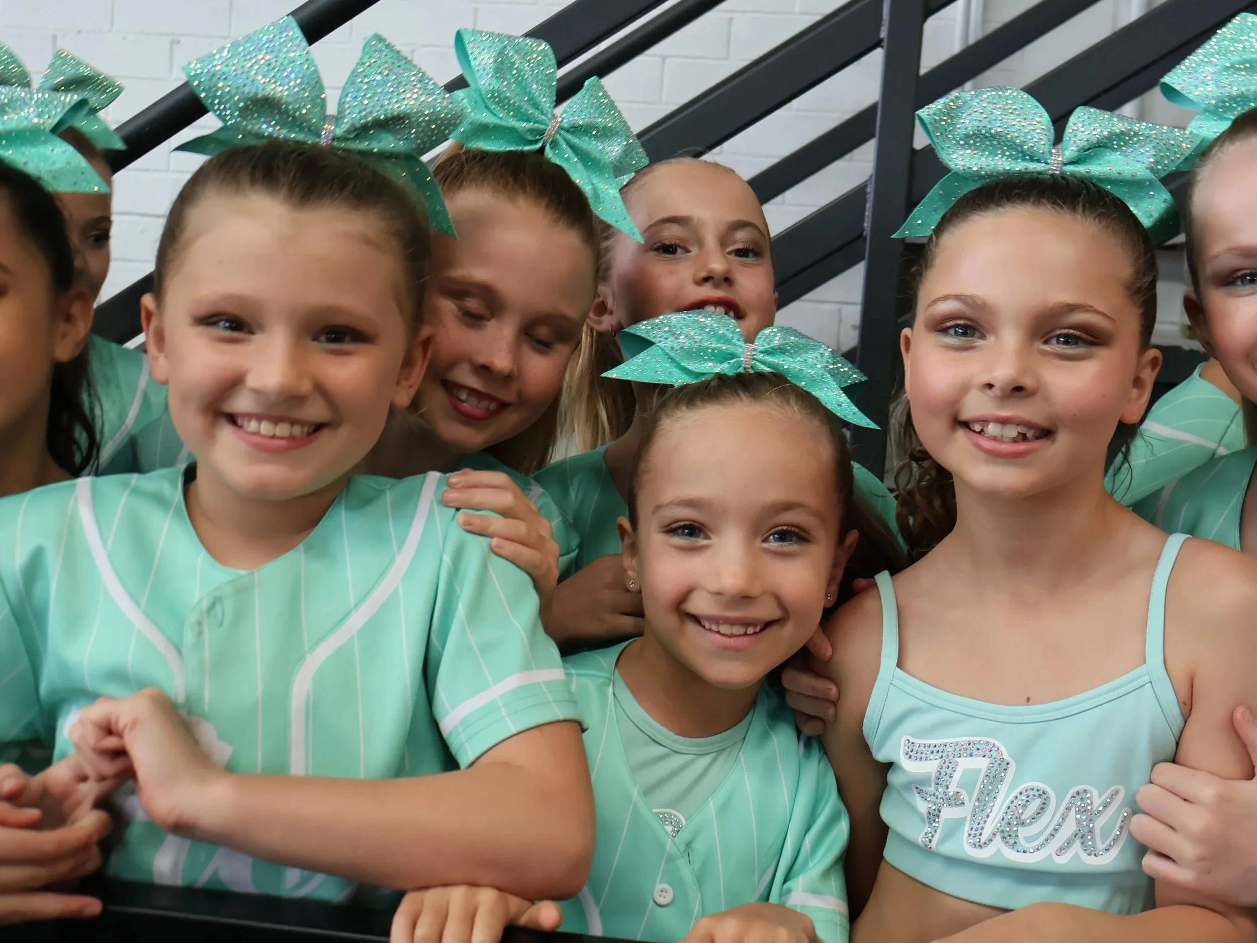 Group of young cheerleaders in matching turquoise uniforms and large bows, smiling at the camera.