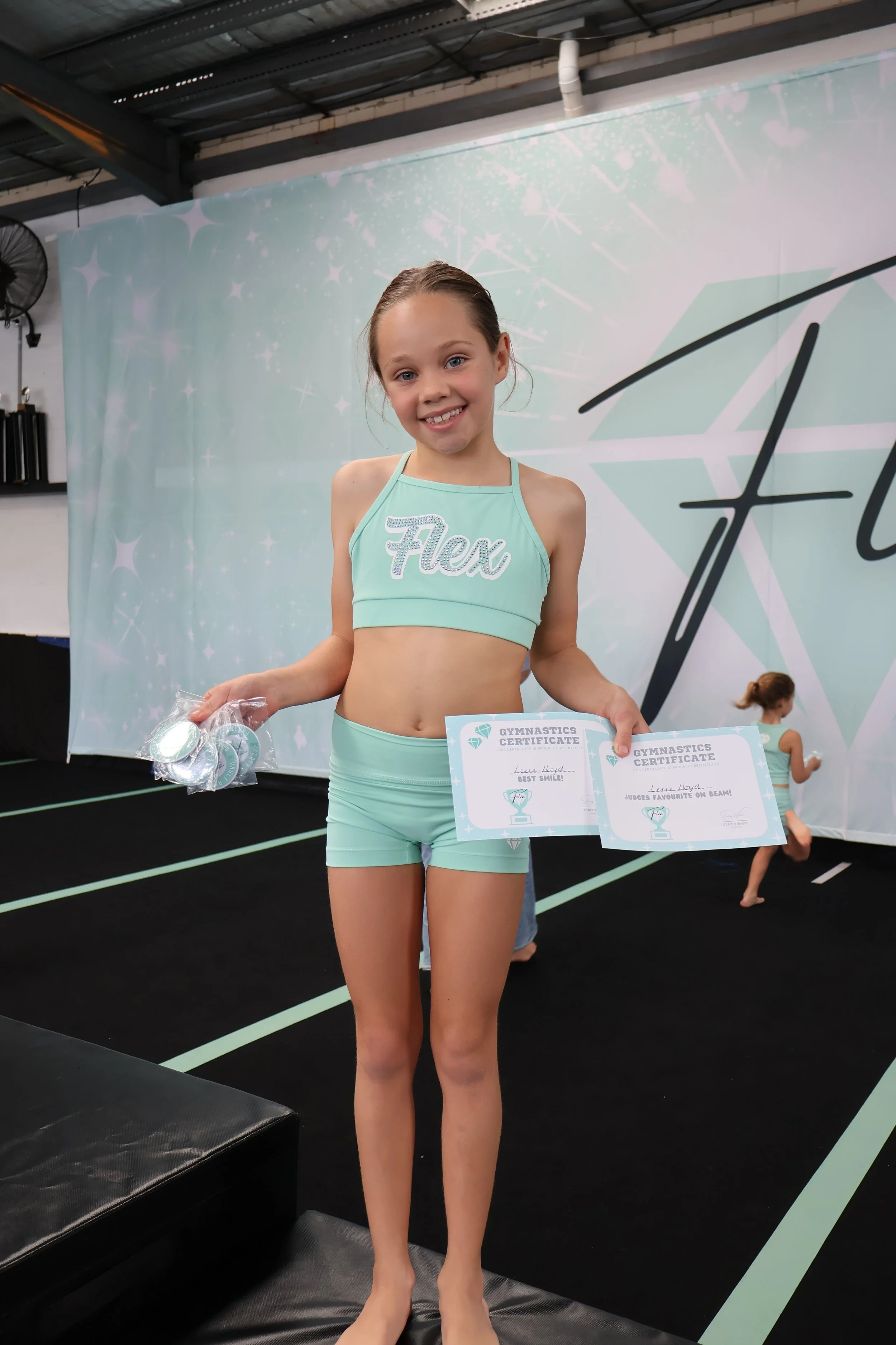 A young girl in a light turquoise gymnastics outfit holding certificates and medals, smiling at a gymnastics event.