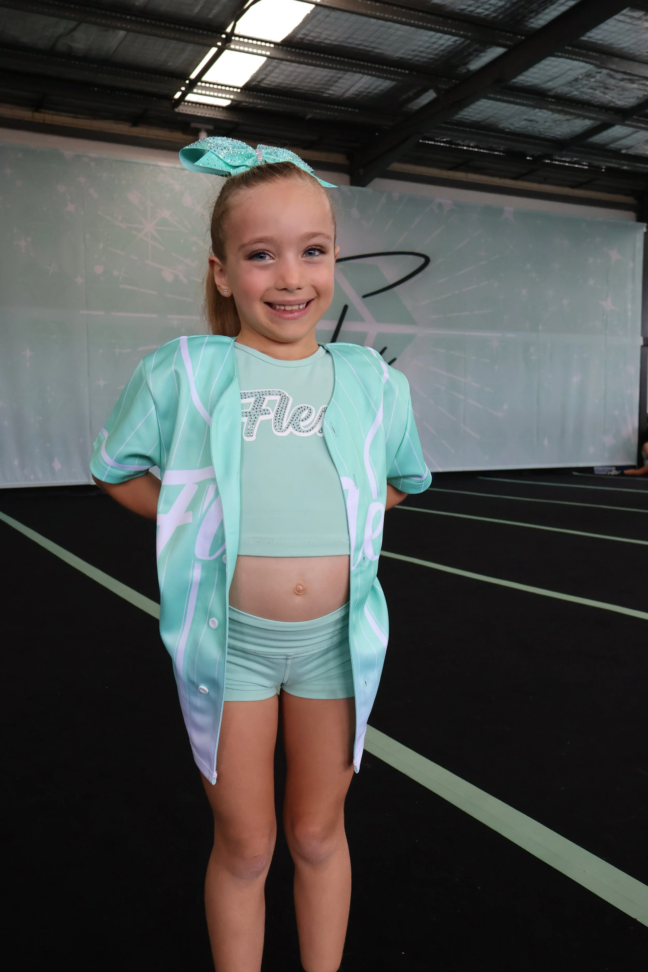 Young girl dressed in a teal cheerleading outfit, smiling, with a teal bow in her hair, standing on a black floor with green lines under a metal roof.