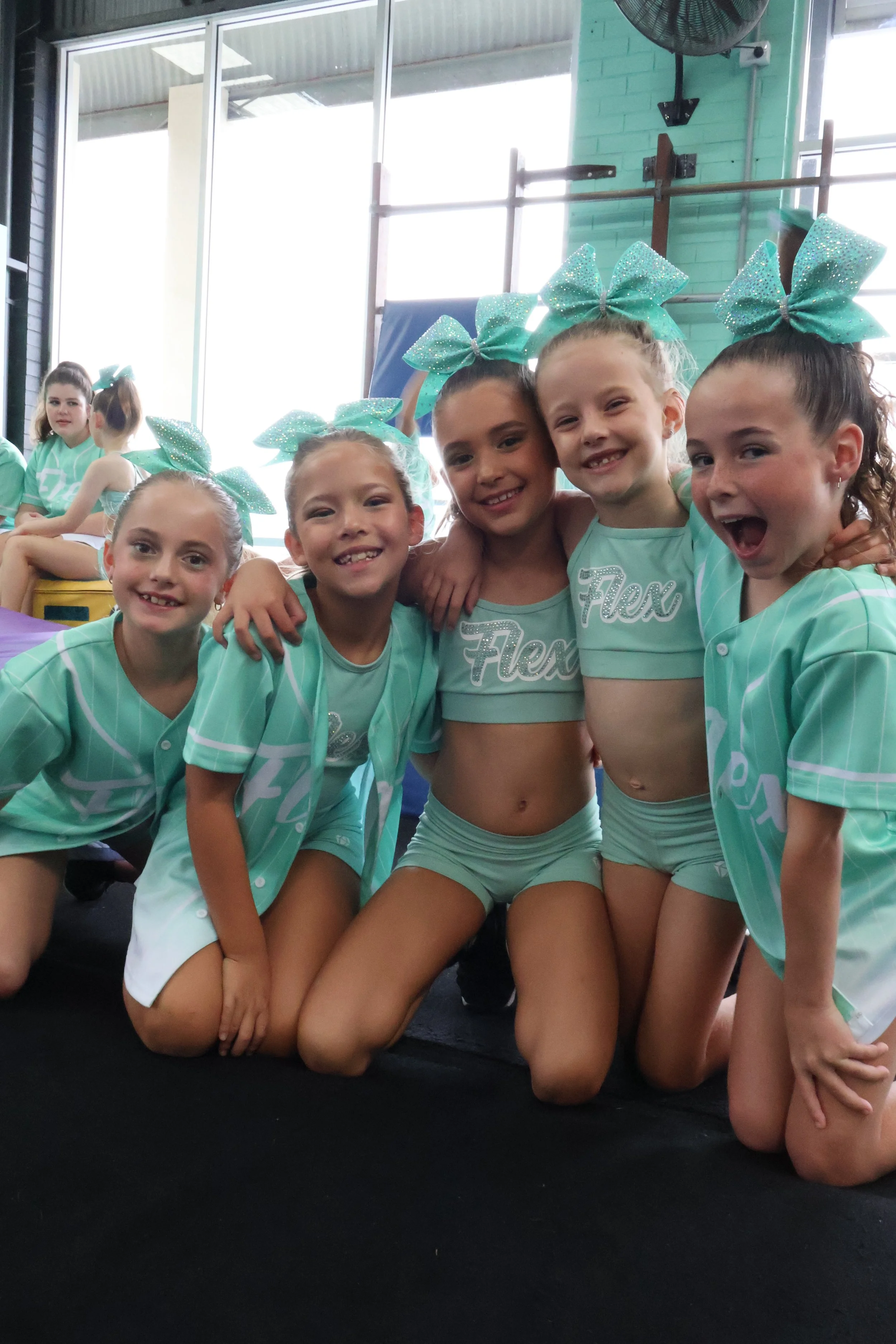 Teenage cheerleaders in teal uniforms and bows posing together for a photo, some kneeling in front with others sitting in the background in a gym.