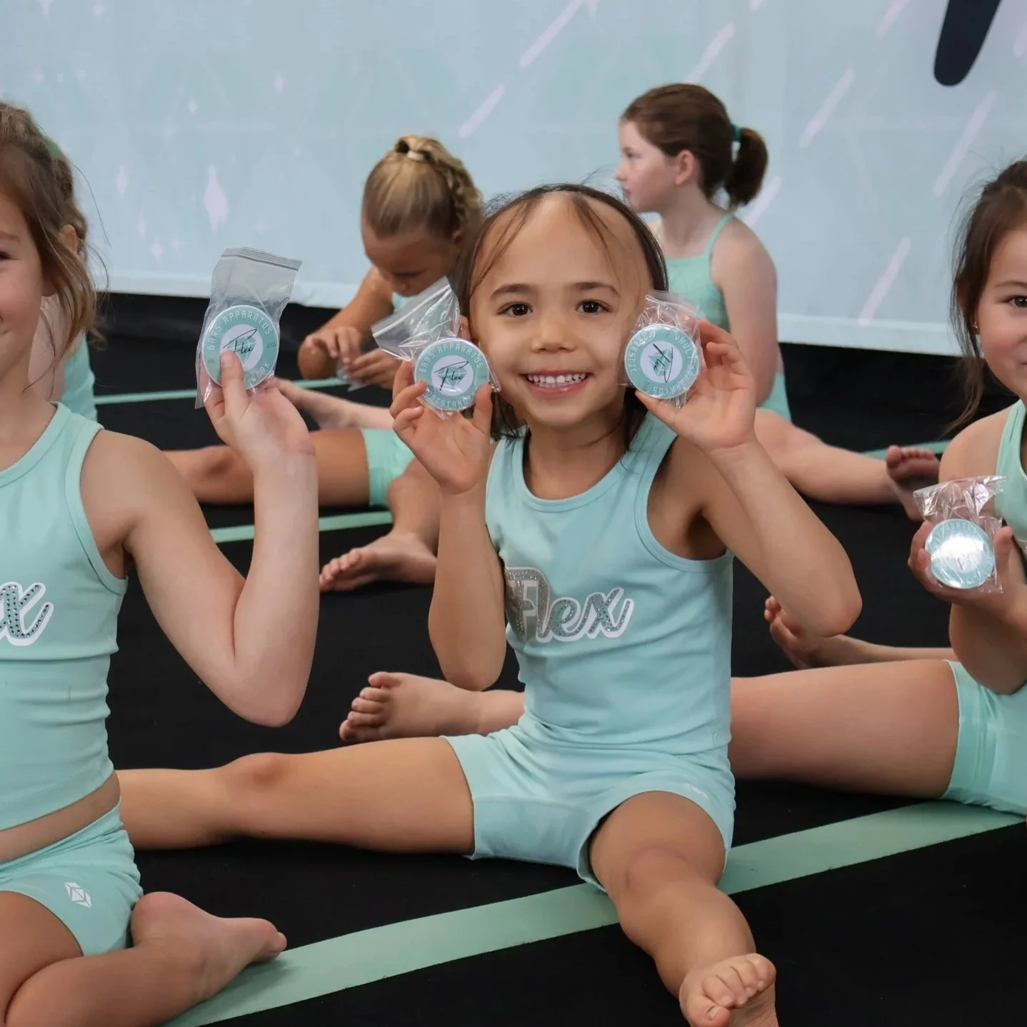 Young girls in matching light blue athletic outfits sitting on a black floor with green lines, holding wrapped medals, smiling, and showing their medals at a gym or dance studio.