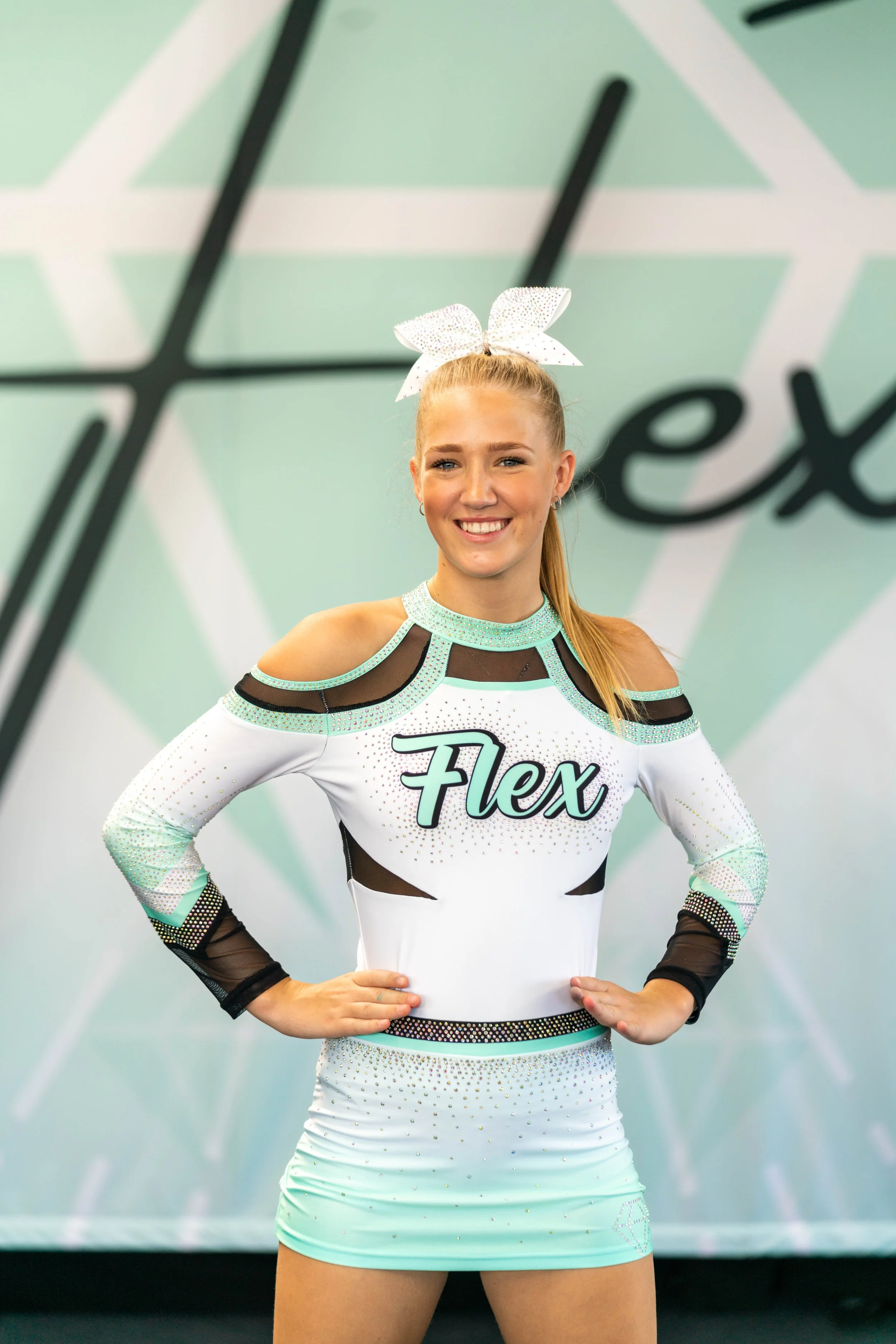 Young female cheerleader smiling at a competition, wearing a white and black cheerleading uniform with turquoise accents and a large white bow in her hair, standing in front of a backdrop that says 'Flex'.