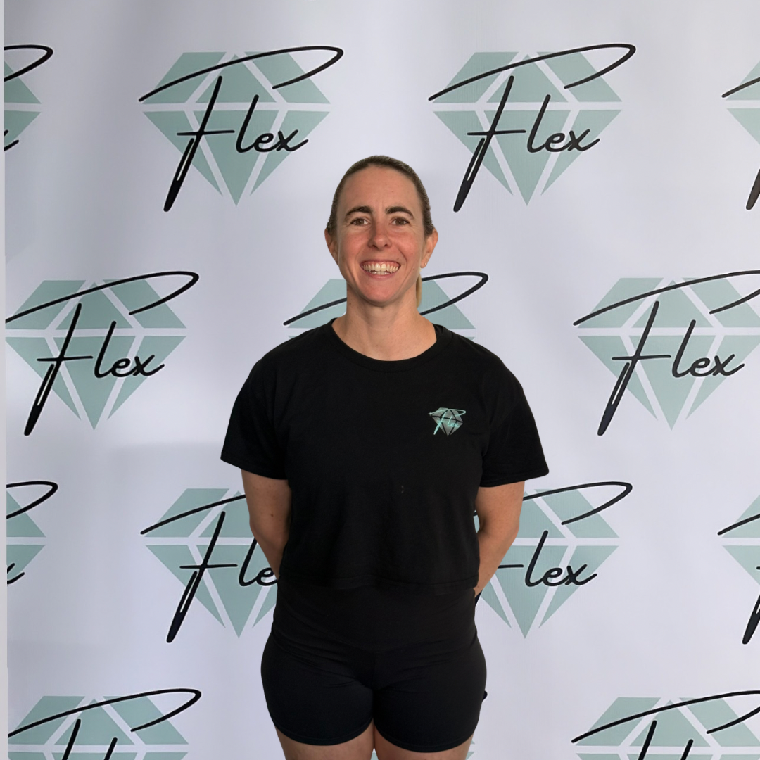 A woman standing in front of a backdrop with a logo that reads 'Flex' and features a stylized diamond. She is smiling, wearing a black T-shirt with the same logo and black shorts.