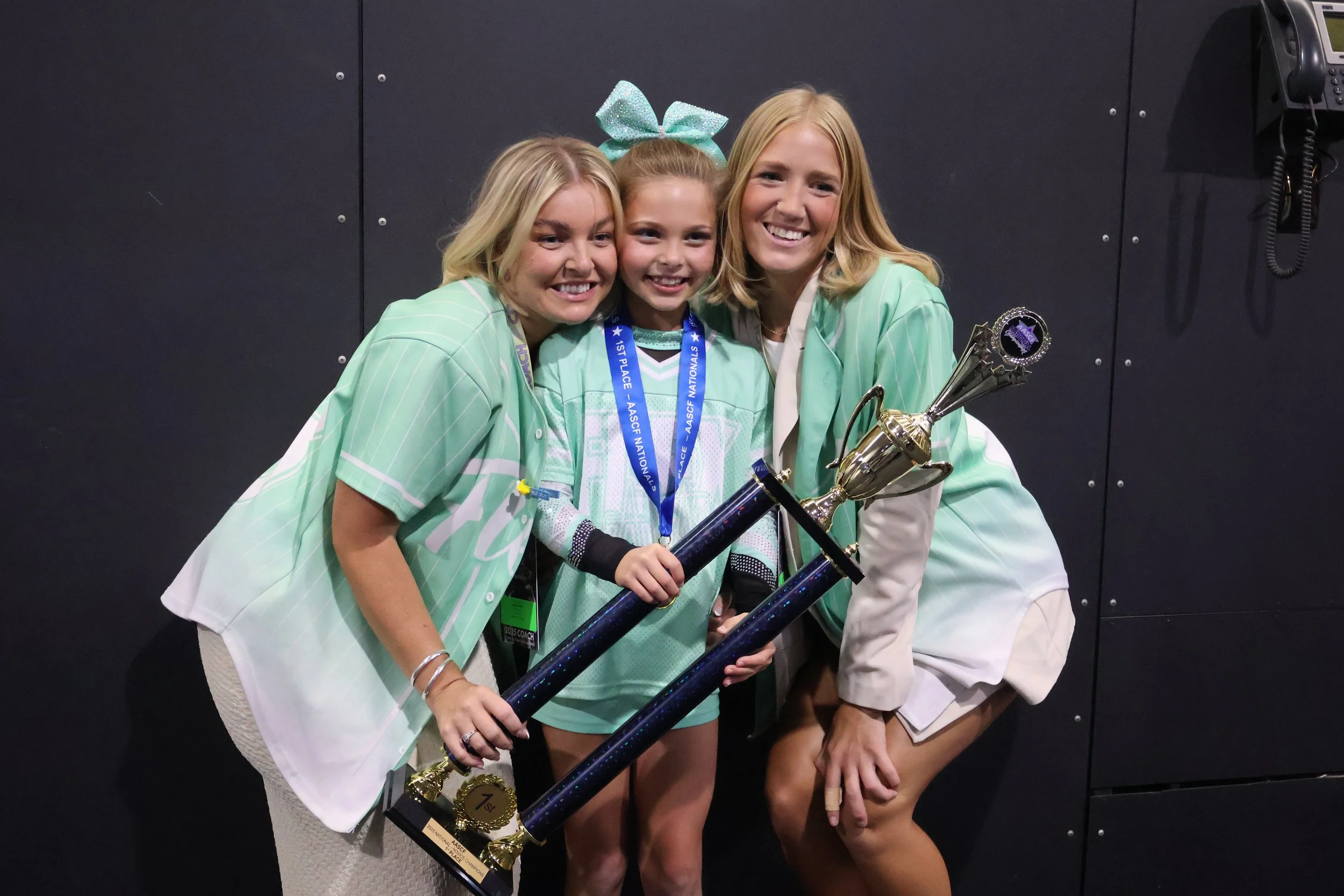 Three smiling females, including a young girl with a light blue medal and a large trophy, posing in front of a black wall.