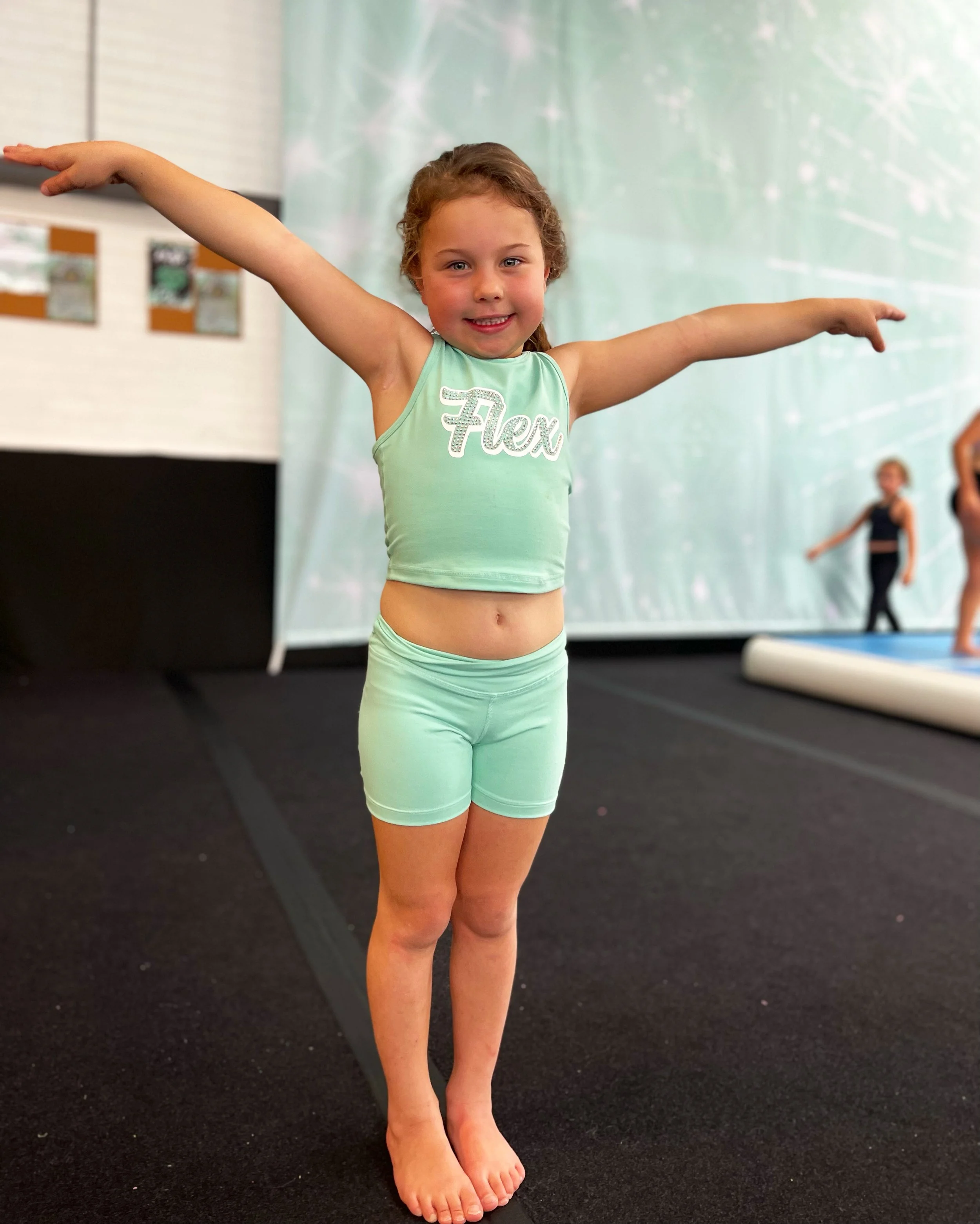 A young girl with curly hair wearing a light green dance outfit with the word 'Flex' on the top, standing with arms outstretched in a dance studio, with other children stretching in the background.
