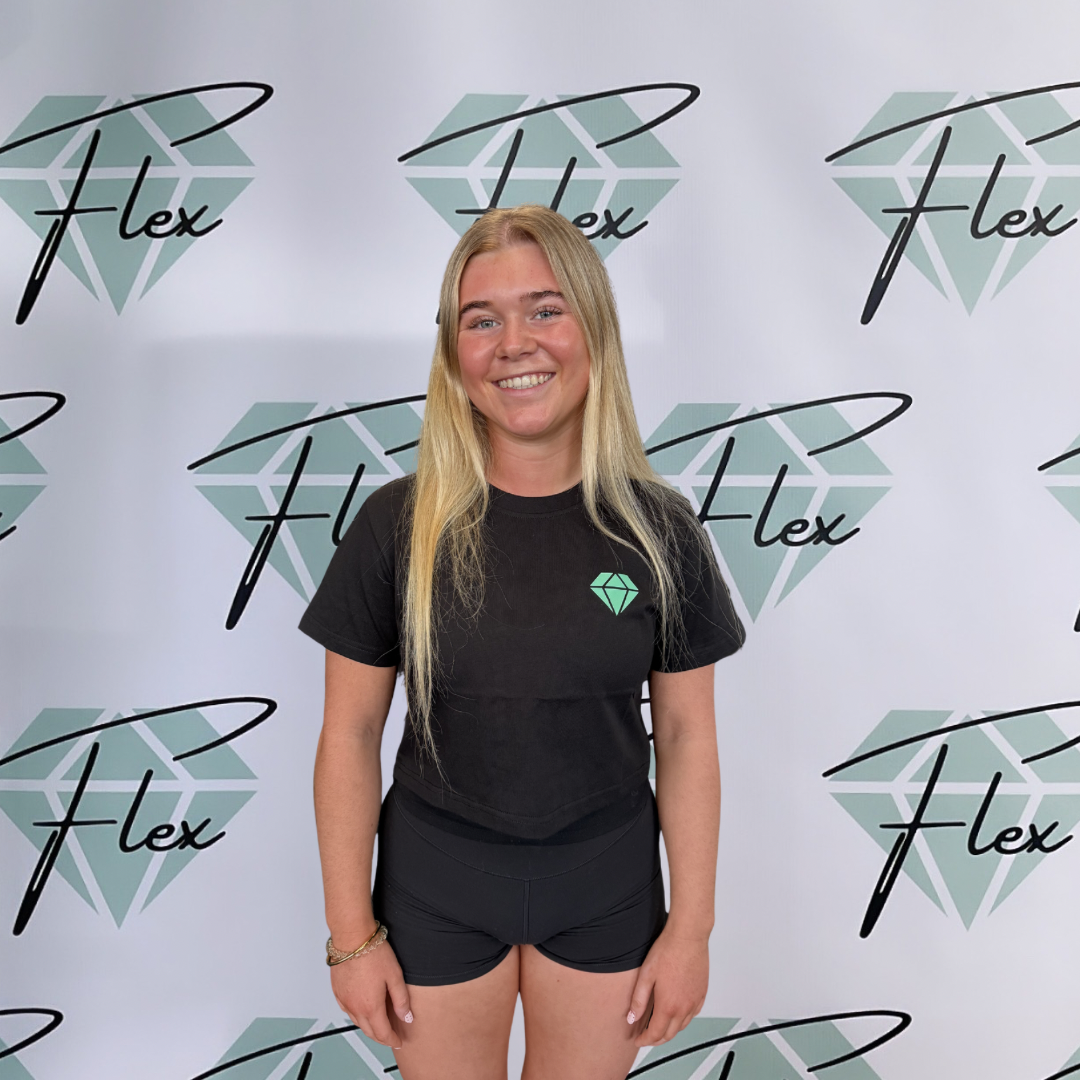 A young woman with long blonde hair smiling at the camera, standing in front of a backdrop with a diamond logo and the word 'Flex' written multiple times.