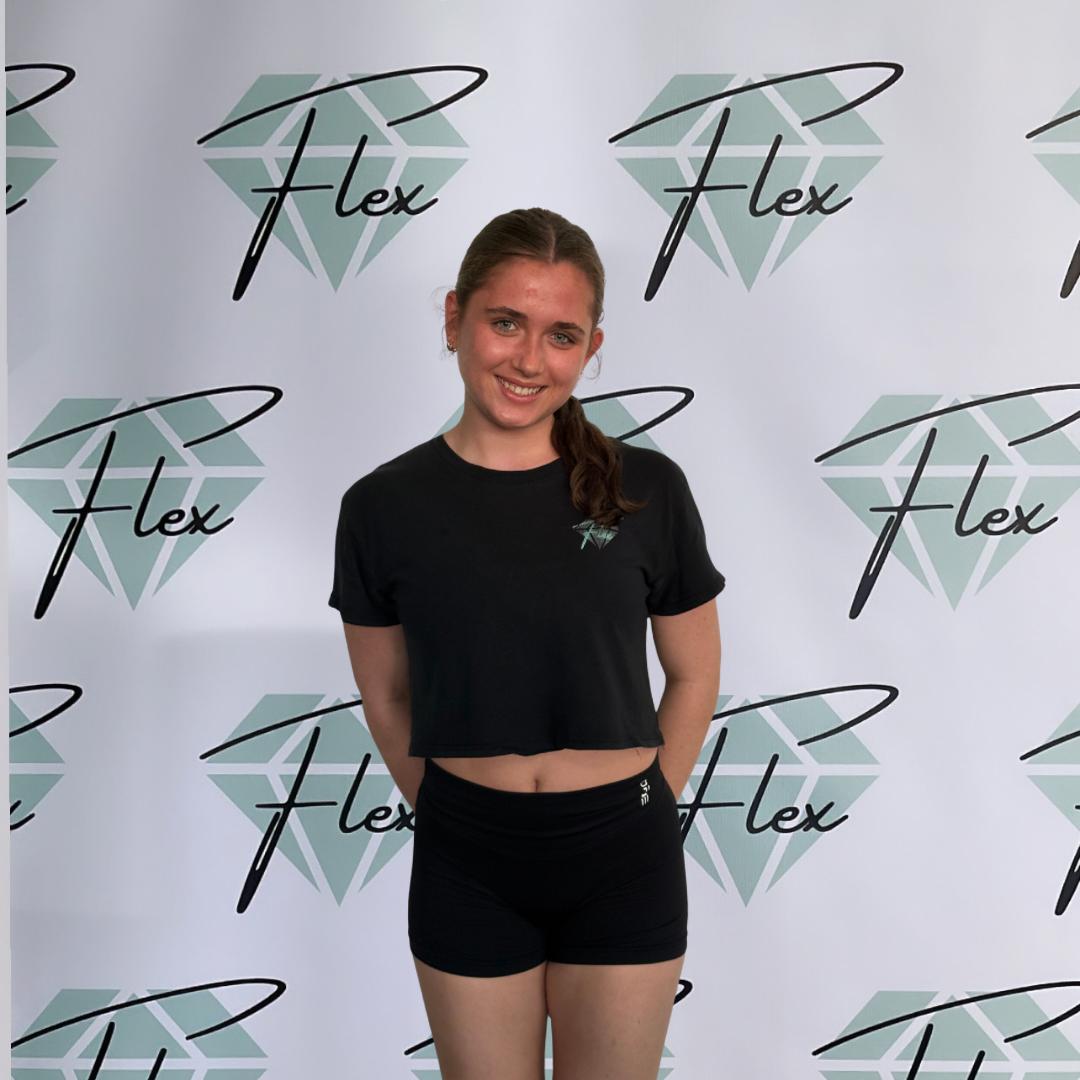 A young woman smiling and standing in front of a backdrop with the word "Flex" and a diamond logo repeated multiple times. She is wearing a black cropped t-shirt and black shorts.