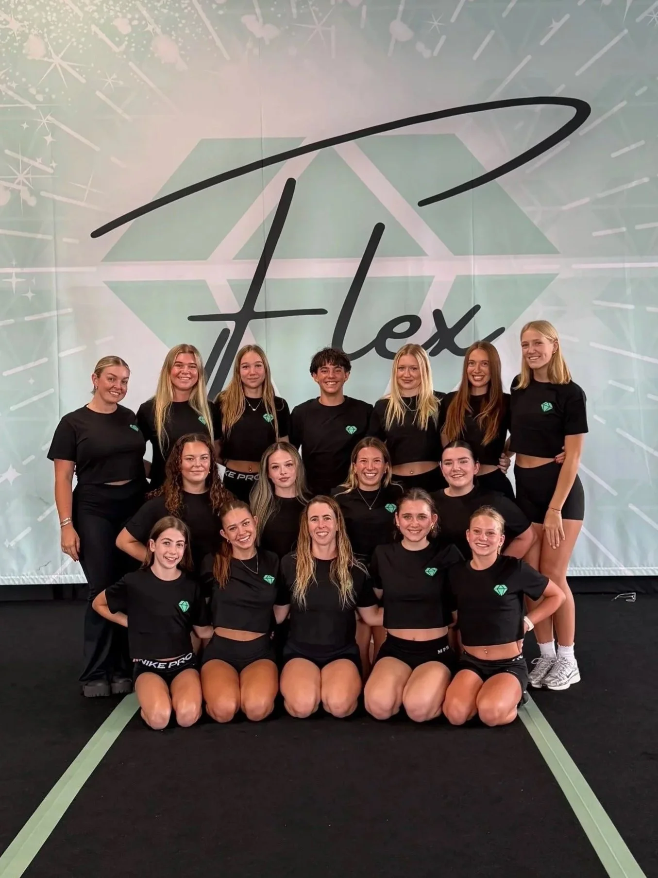 Group of young women in black athletic outfits posing on a stage with a large logo behind them.