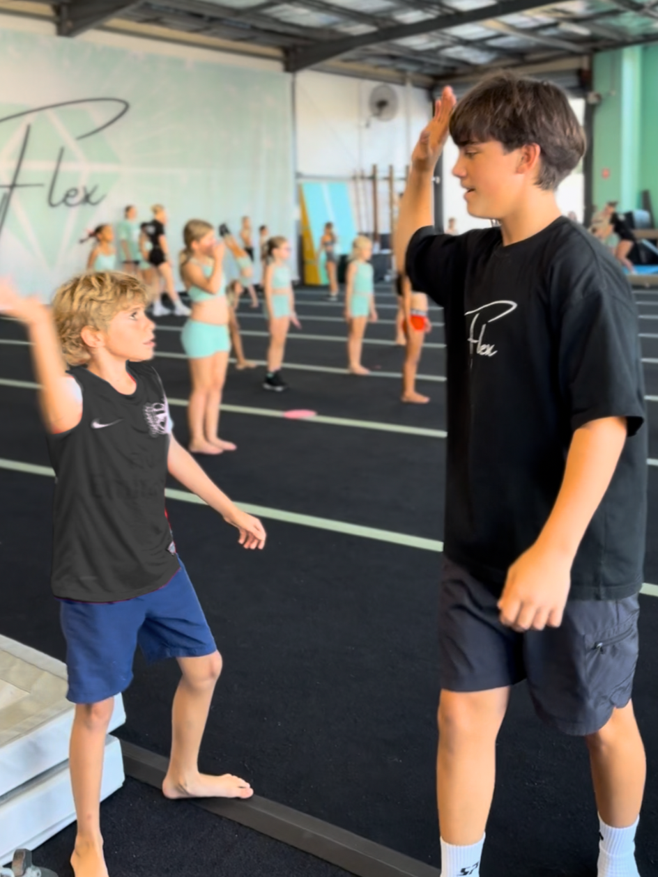 A group of children practicing cheerleading or dance in a gym, with two boys in the foreground. One boy is giving a high five to the other, who is smiling. Other children are lined up in the background.