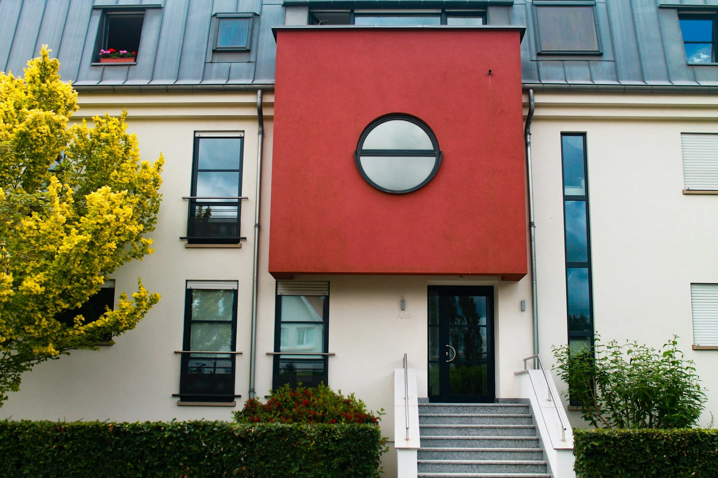 Modern multi-story residential building with a prominent red facade and a circular window, surrounded by stairs, greenery, and yellow trees.
