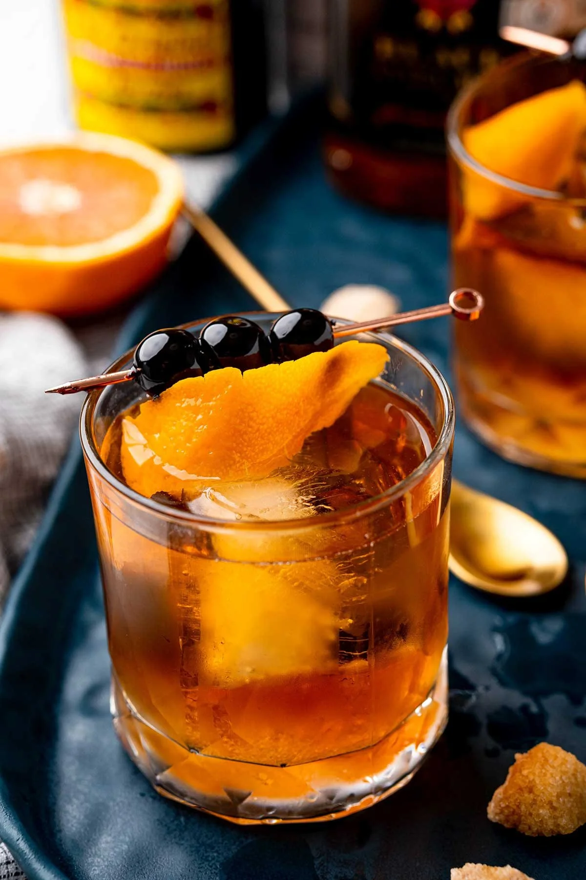 A glass of iced tea garnished with a slice of orange and three black cherries, with a halved orange in the background and a spoon with a lemon wedge on a dark tray.