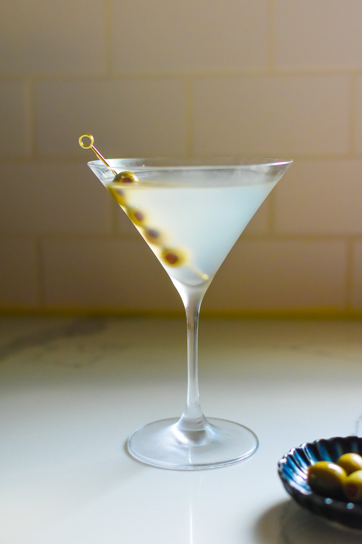A martini glass filled with a white cocktail, garnished with skewered green and black olives, placed on a light-colored surface with a neutral background.