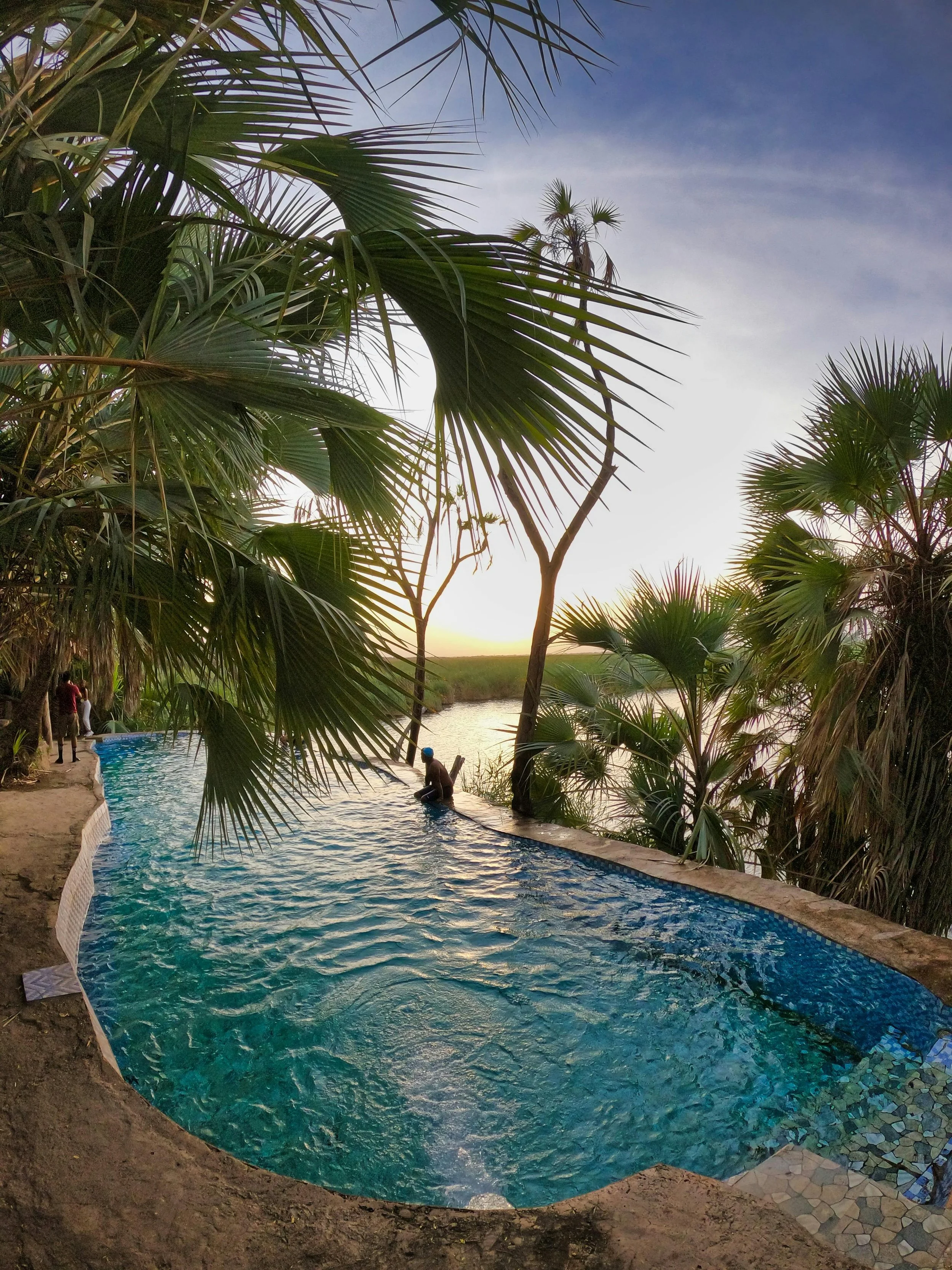 A natural swimming pool surrounded by lush tropical trees with the sun setting in the background.