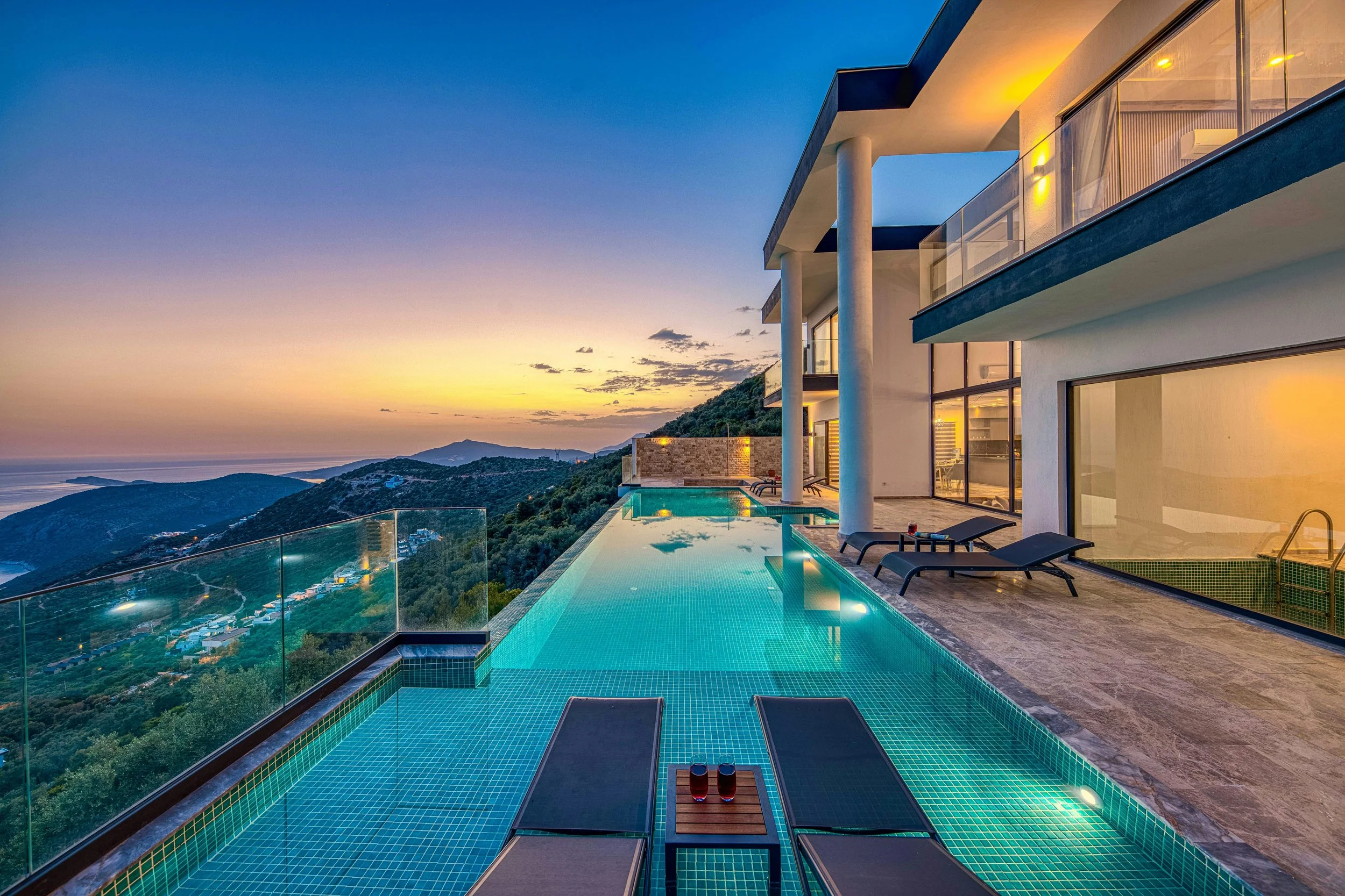 Luxury modern house with a rooftop infinity pool overlooking a mountainous landscape at sunset, with lounge chairs and glass railing.