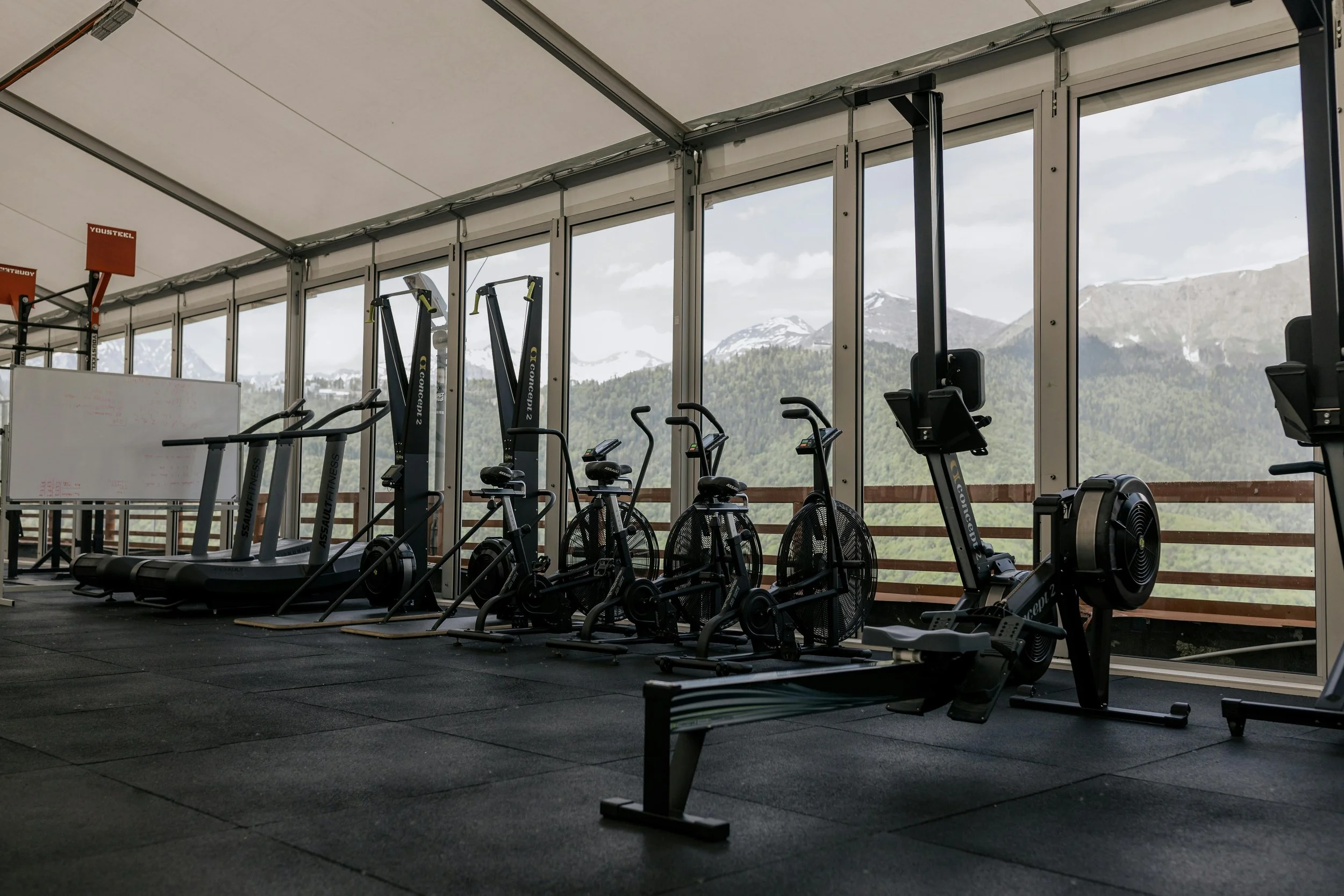 A gym with treadmills, exercise bikes, and ellipticals inside a room with large windows showing mountains in the background.