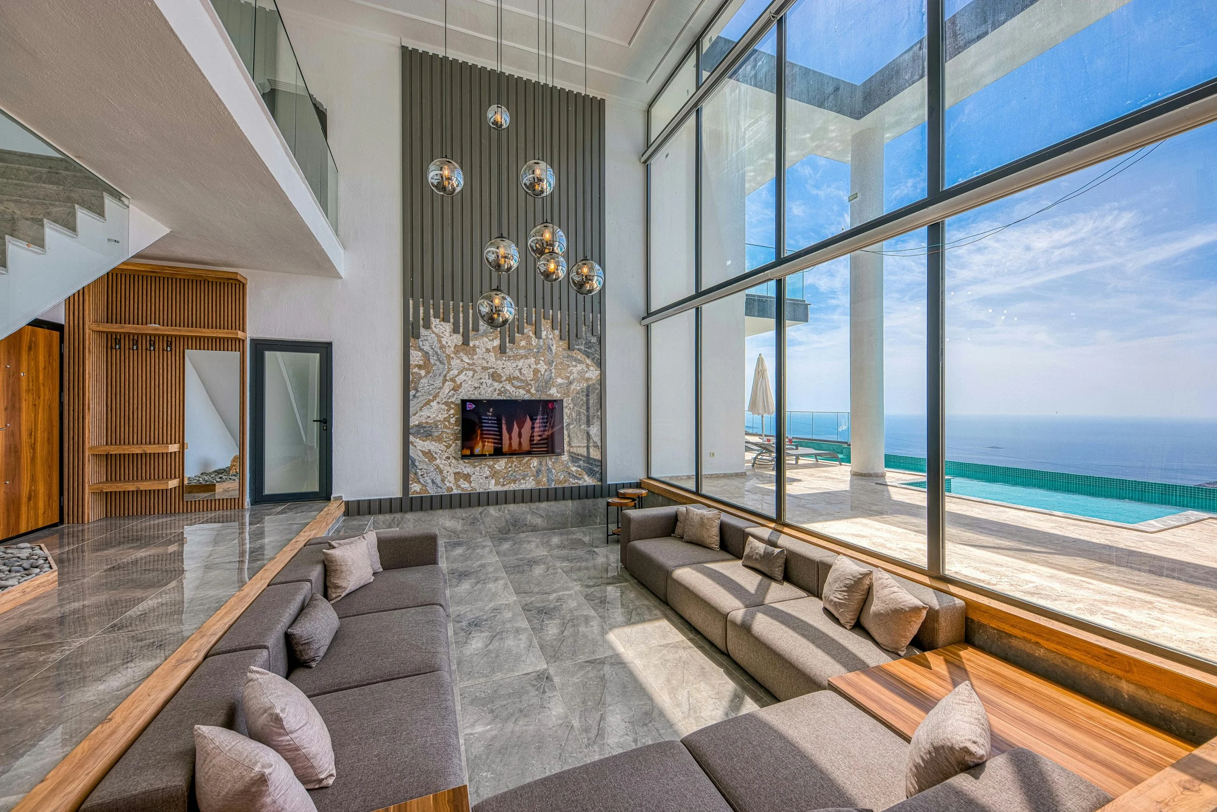 Modern living room with large floor-to-ceiling windows overlooking a swimming pool and ocean, featuring a gray sectional sofa with pillows, a stone wall with a mounted TV, and a decorative chandelier of hanging glass globes.
