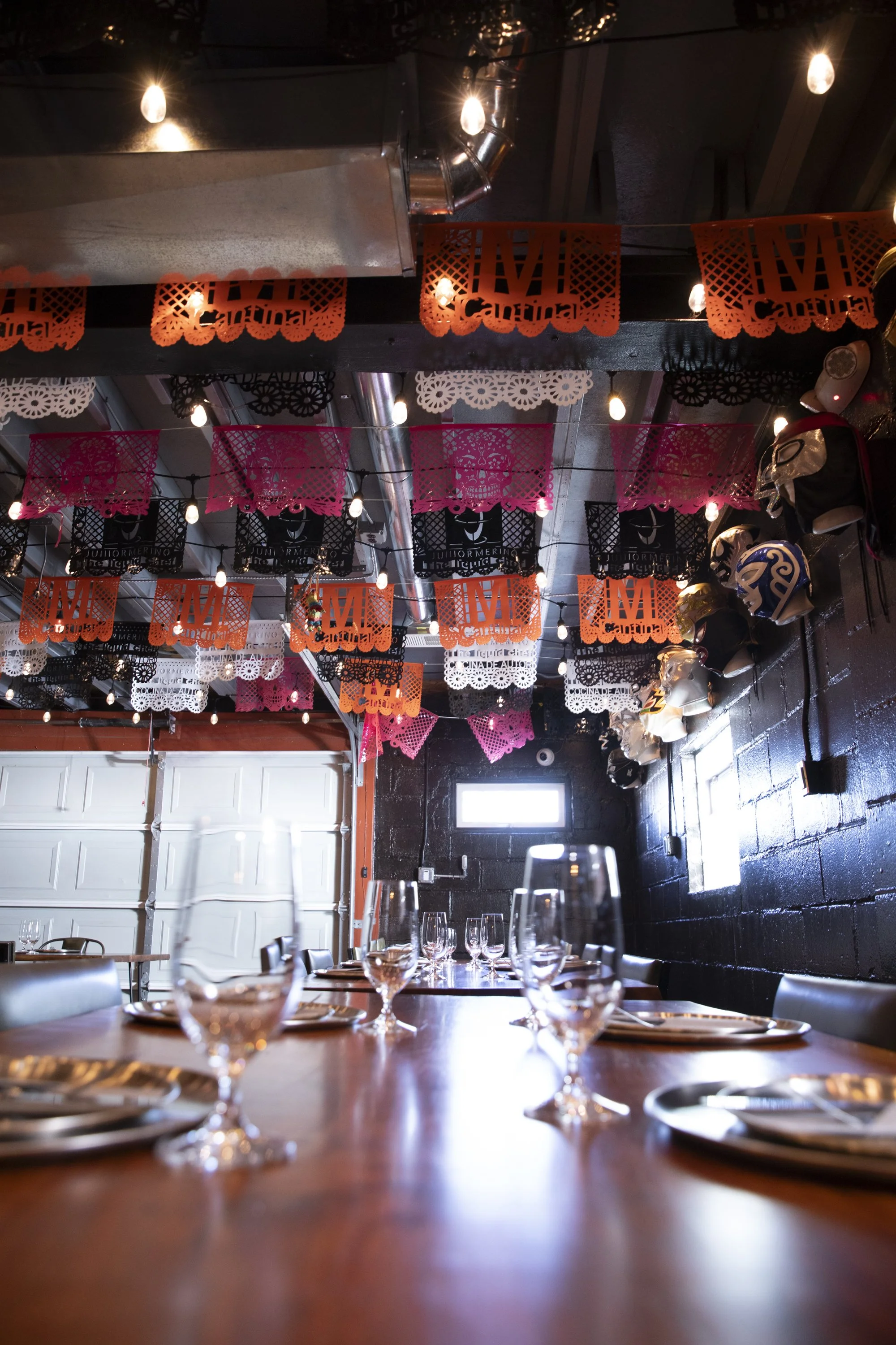 Decorated dining area with colorful papel picado banners hanging from the ceiling, a long wooden table set with wine glasses, stacks of plates, and cutlery, with a window and white garage door visible in the background, indicative of a festive Mexican celebration.