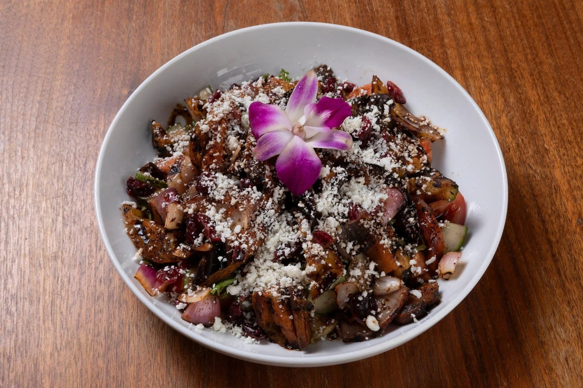 A white bowl of salad topped with shredded cheese and a pink and white orchid flower on top, placed on a wooden table.