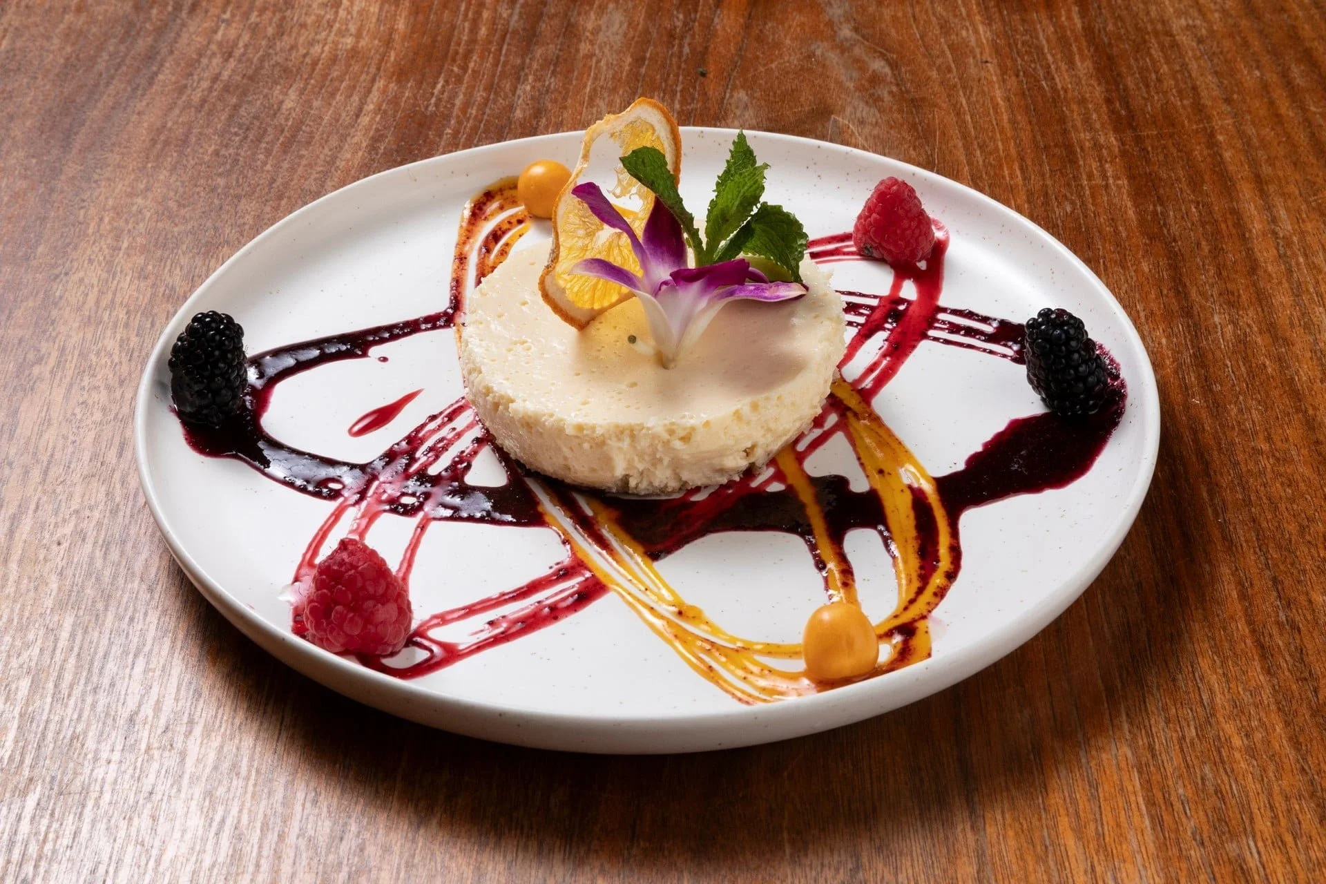 A white plate with a cheesecake garnished with edible flowers, mint leaves, and various fruits, drizzled with berry, caramel, and chocolate sauces on a wooden surface.