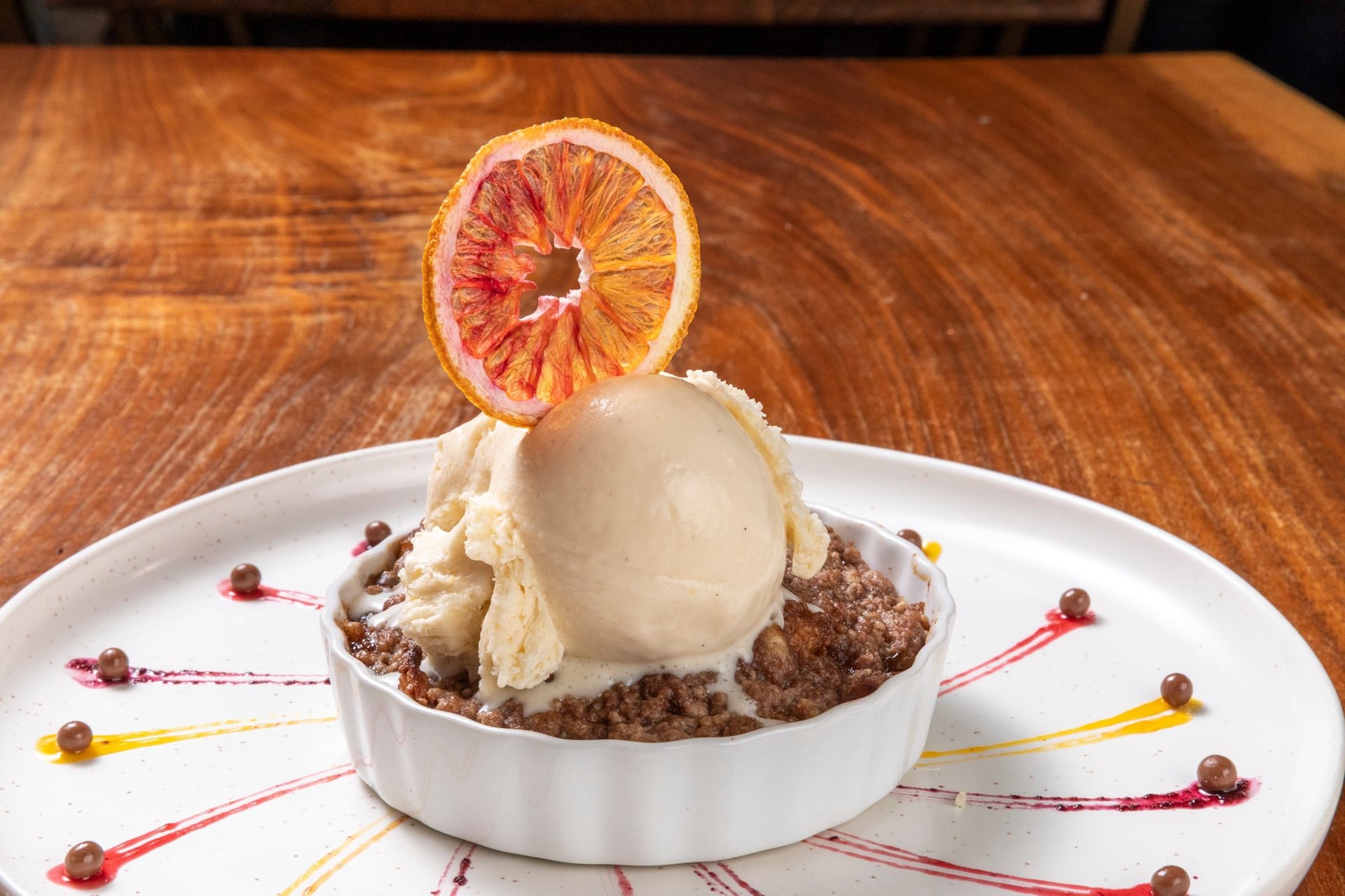 Dessert dish with vanilla ice cream, topped with a dried orange slice, served in a white ramekin on a decorated white plate.