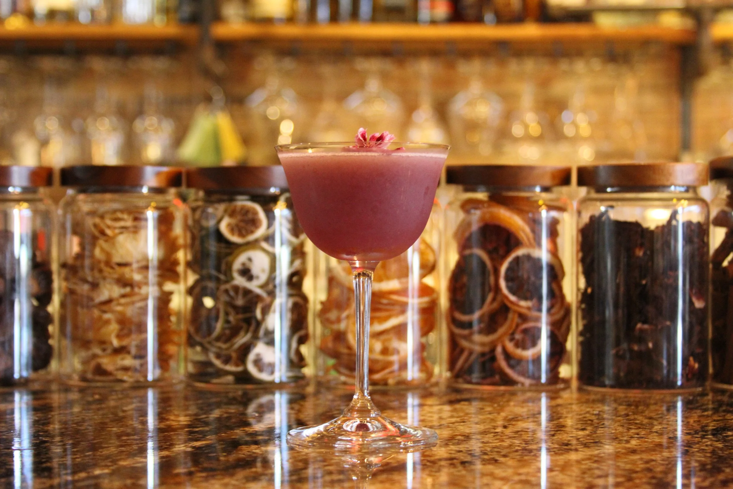 A pink cocktail garnished with small pink flowers on top, placed on a wooden bar counter with glass jars filled with dried herbs and fruit slices in the background.