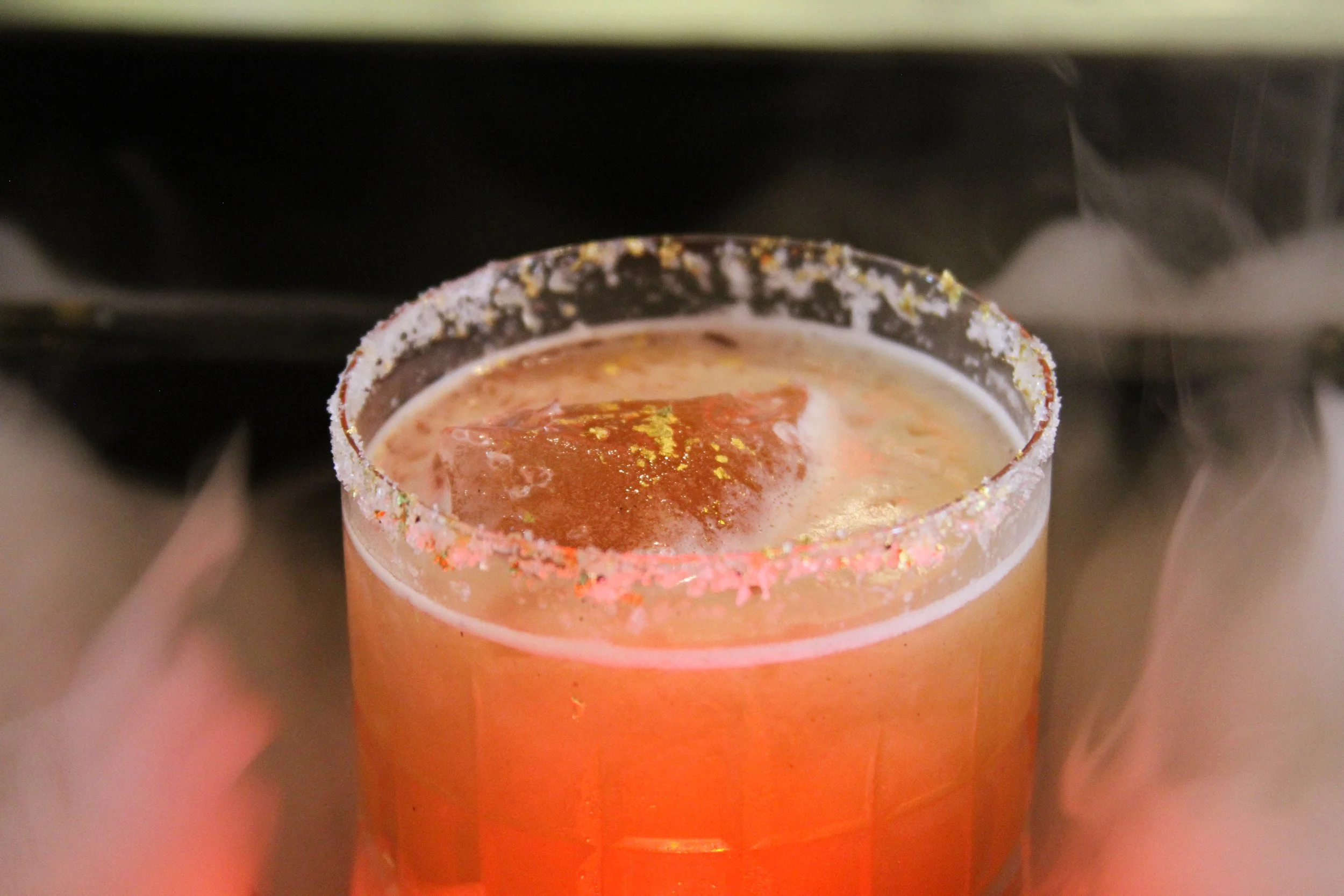 Close-up of a festive cocktail glass with a salted rim, garnished with gold flakes and a red cherry or fruit piece, on a blurred background.