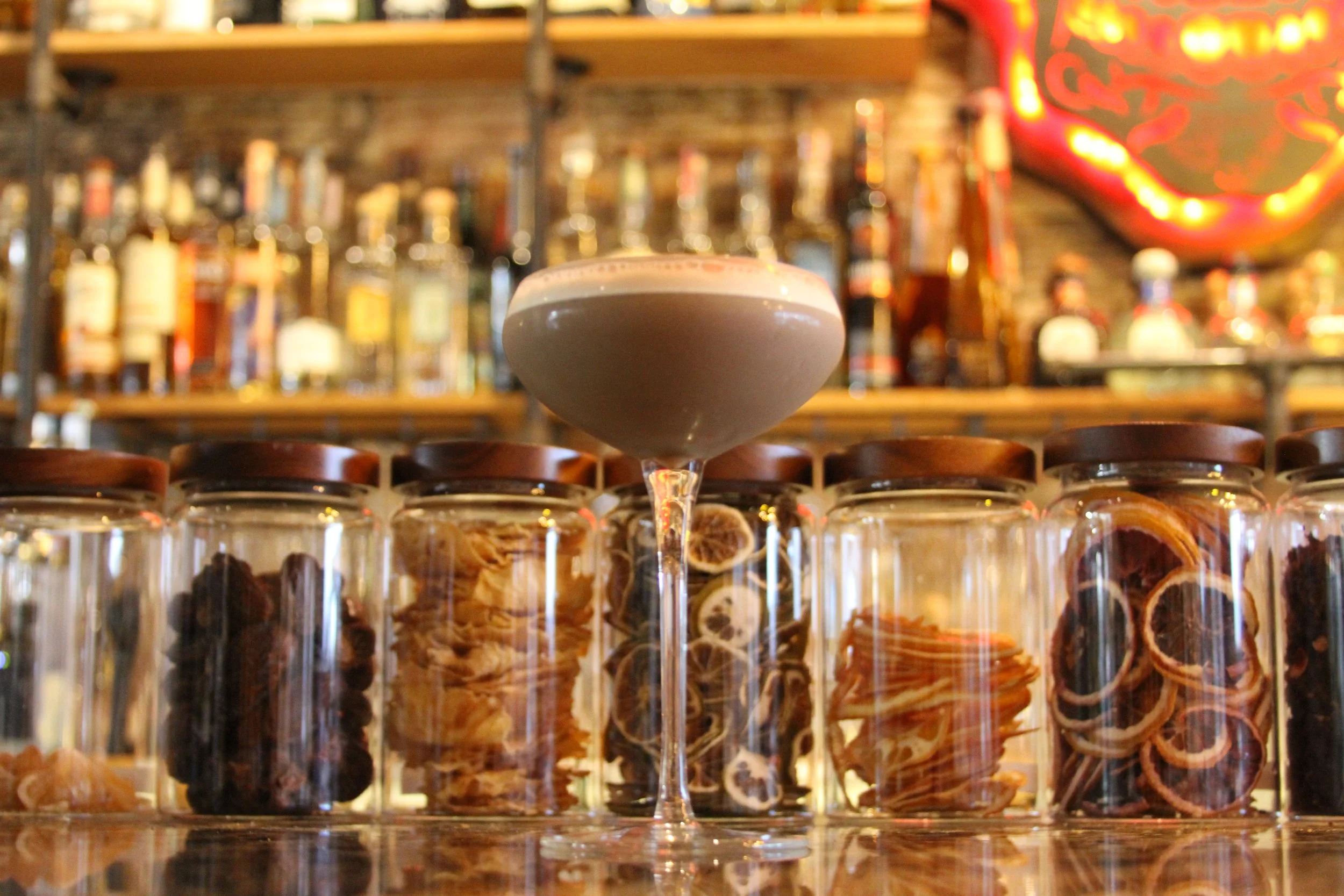 A cocktail glass filled with a dark beverage topped with whipped cream, placed in front of a bar with various bottles of alcohol and a neon sign in the background.