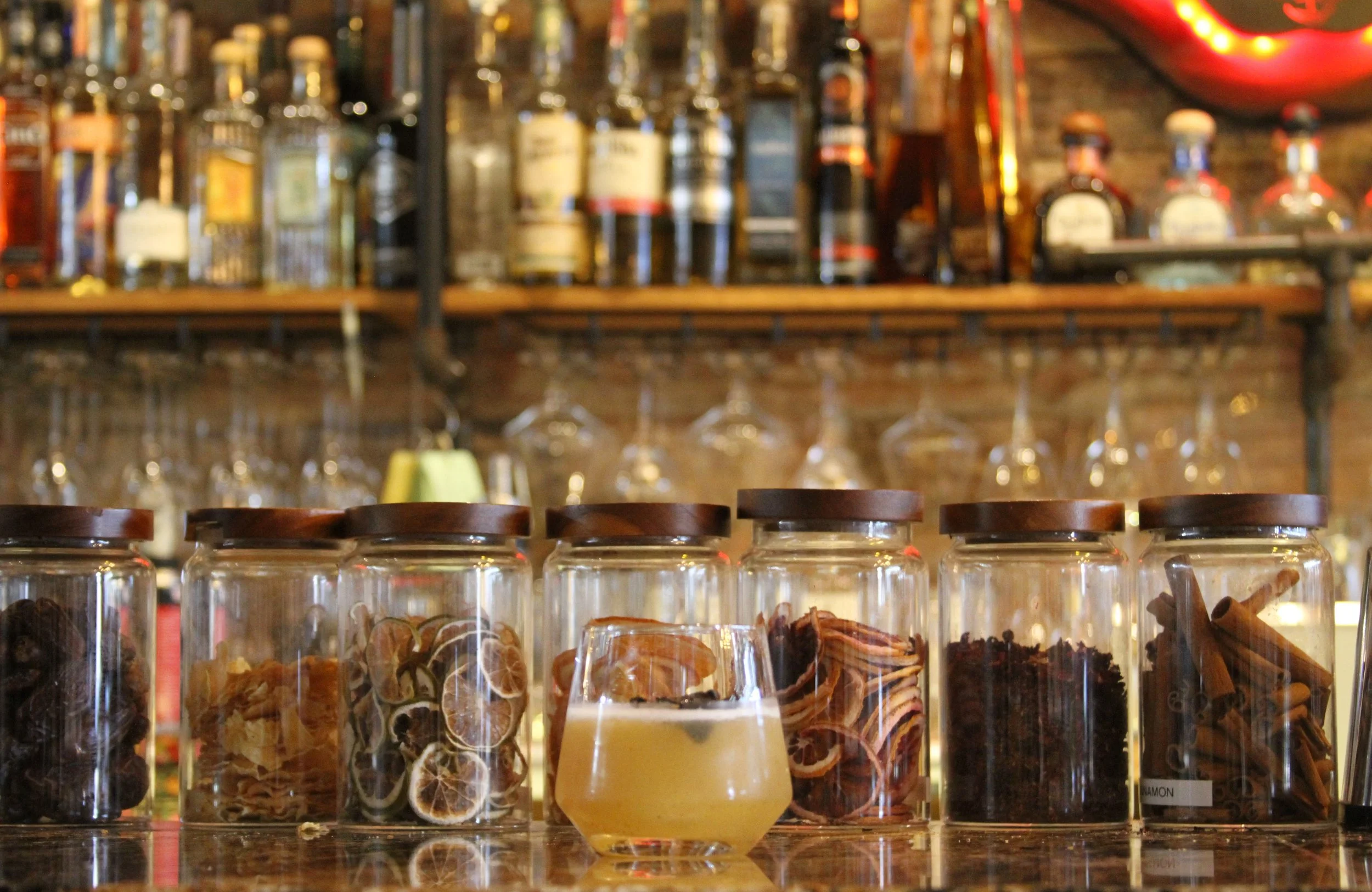 A row of clear jars with dark brown wooden lids containing various dried herbs and spices, placed on a display shelf in a bar or café setting, with bottles of alcohol and shot glasses in the background.