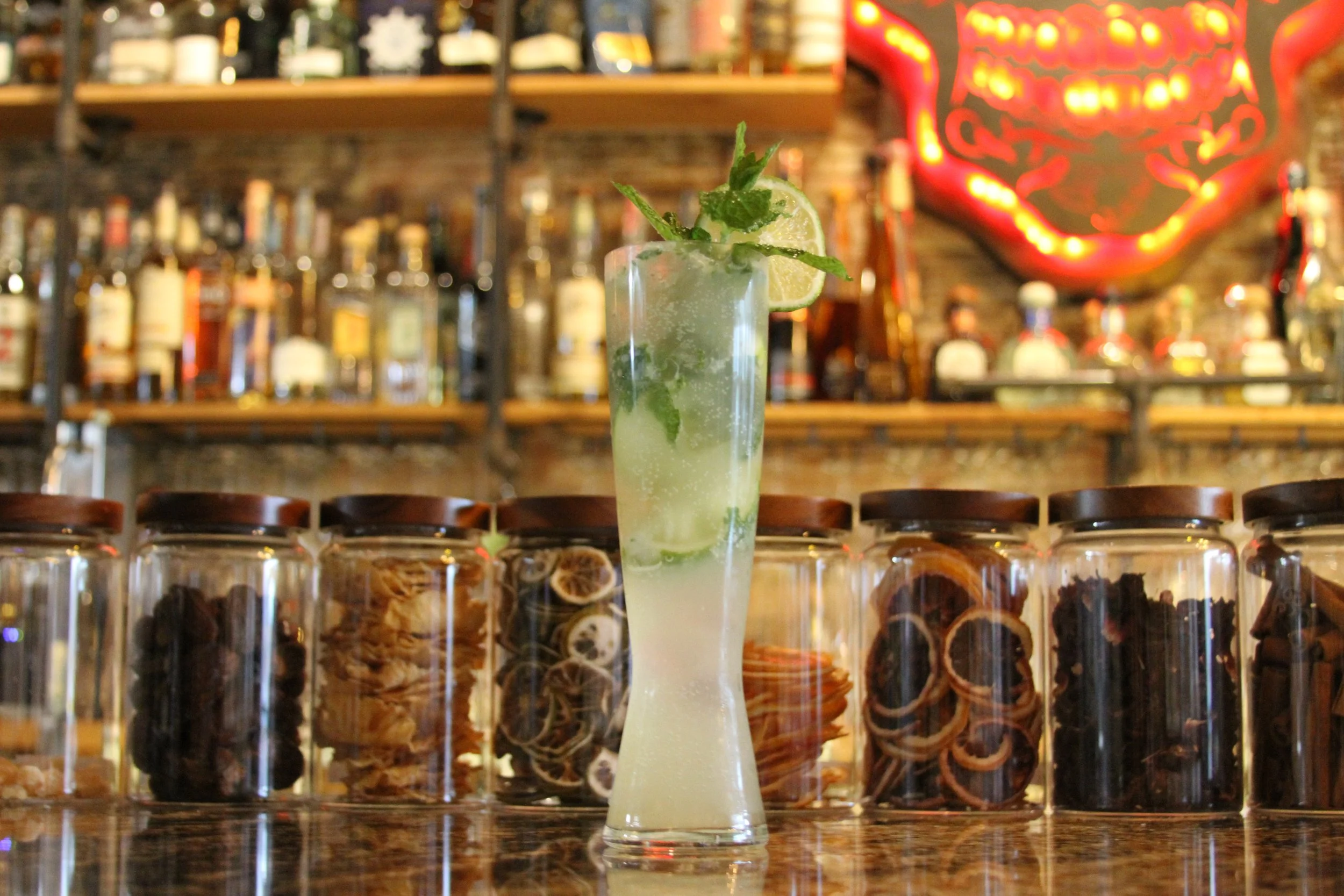 A tall glass of mojito with lime slices and mint leaves, set on a bar counter with blurred bottles in the background and a neon sign on the wall.