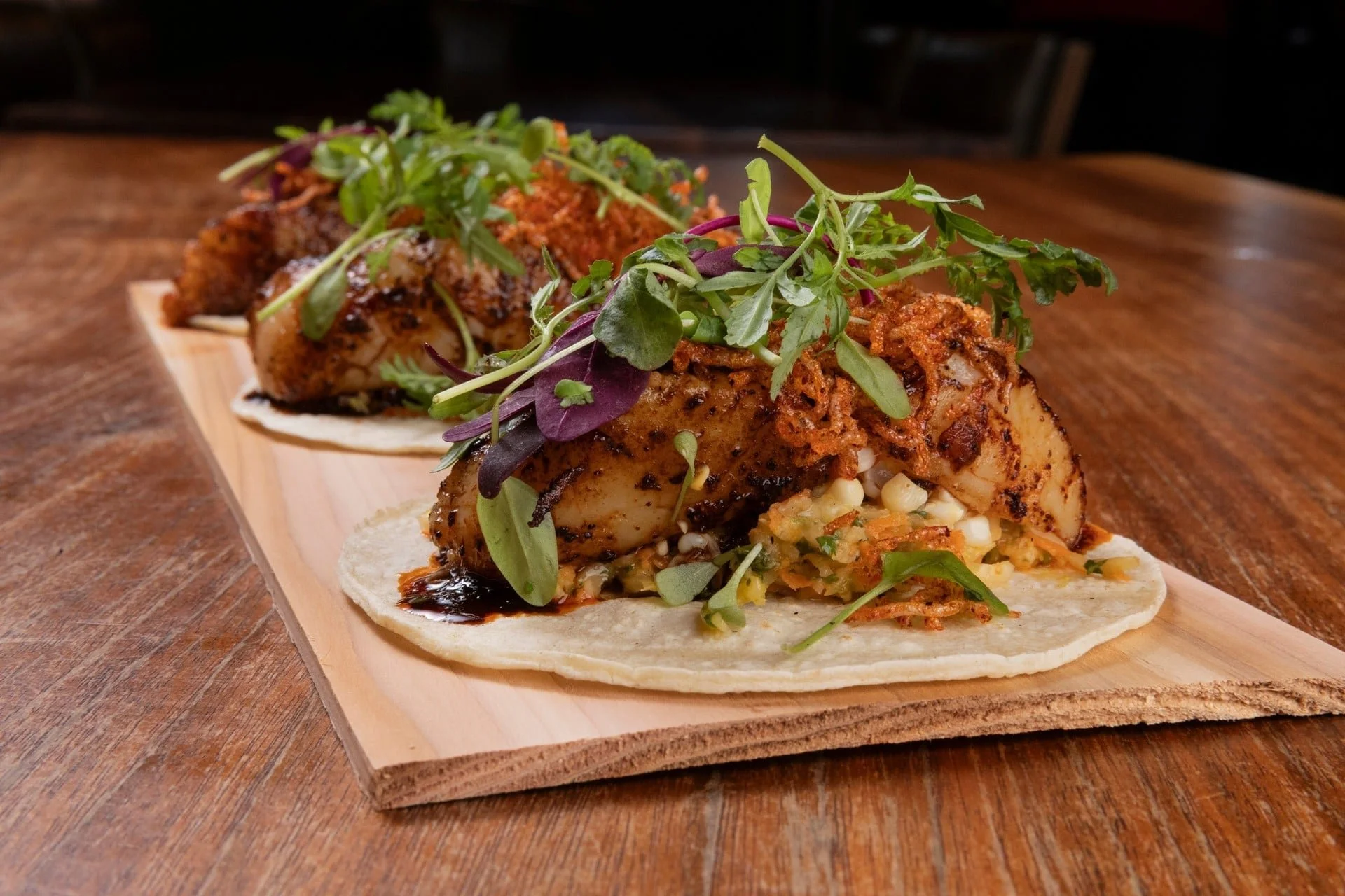 Close-up of two tacos filled with grilled chicken, topped with microgreens, on a wooden serving board.