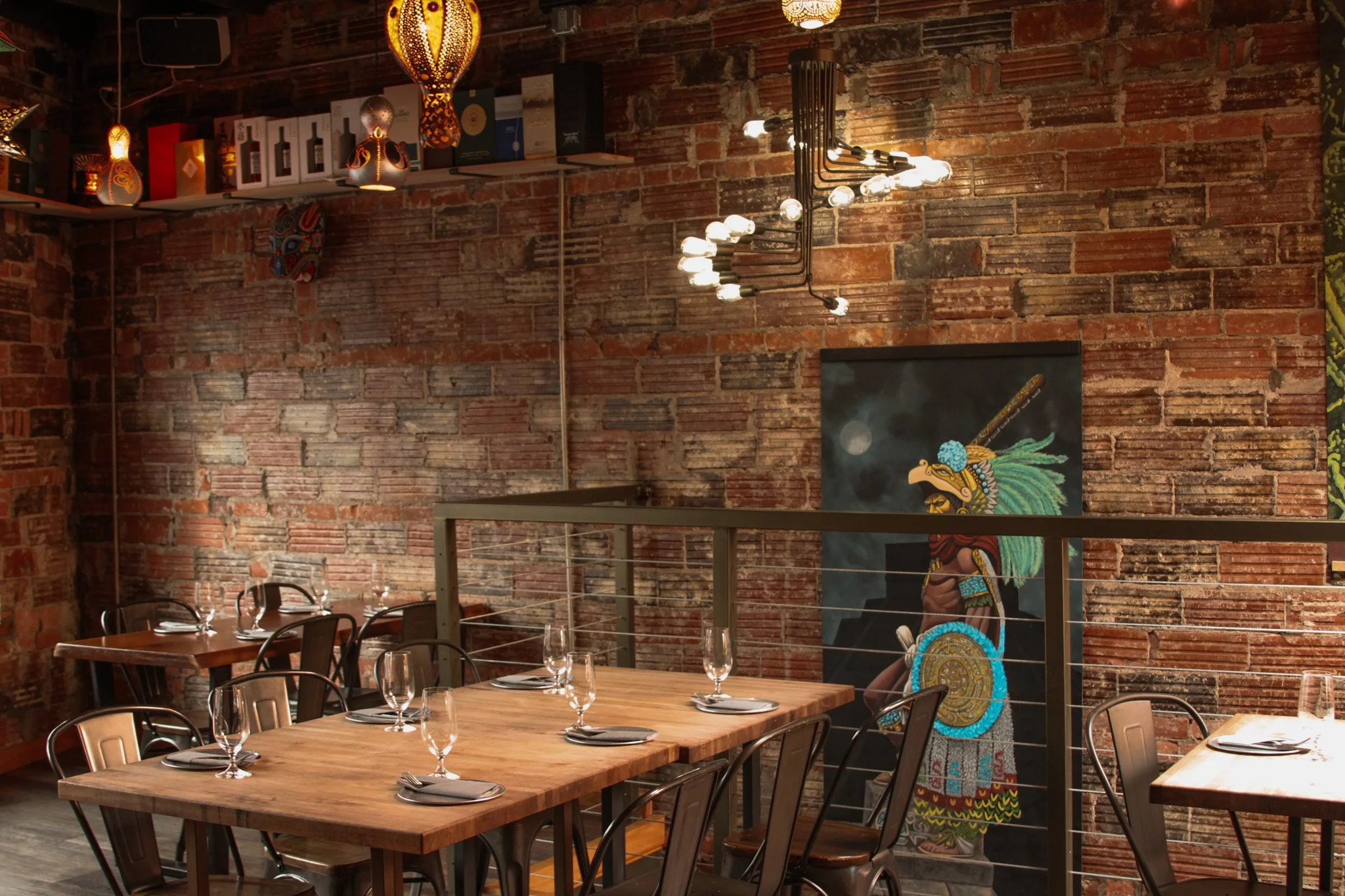 Interior of a restaurant with brick walls, rectangular wooden tables set with wine glasses and napkins, black chairs, decorative hanging lights, and artwork including a colorful depiction of a Maya warrior.