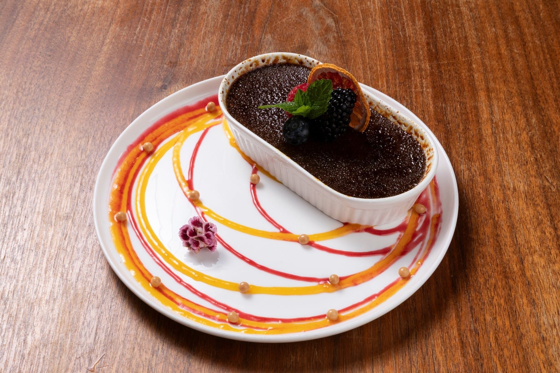 Chocolate dessert in a white ramekin topped with blackberries, a slice of orange, and a mint leaf, served on a decorated white plate with colorful sauce swirls and pearls, on a wooden table.