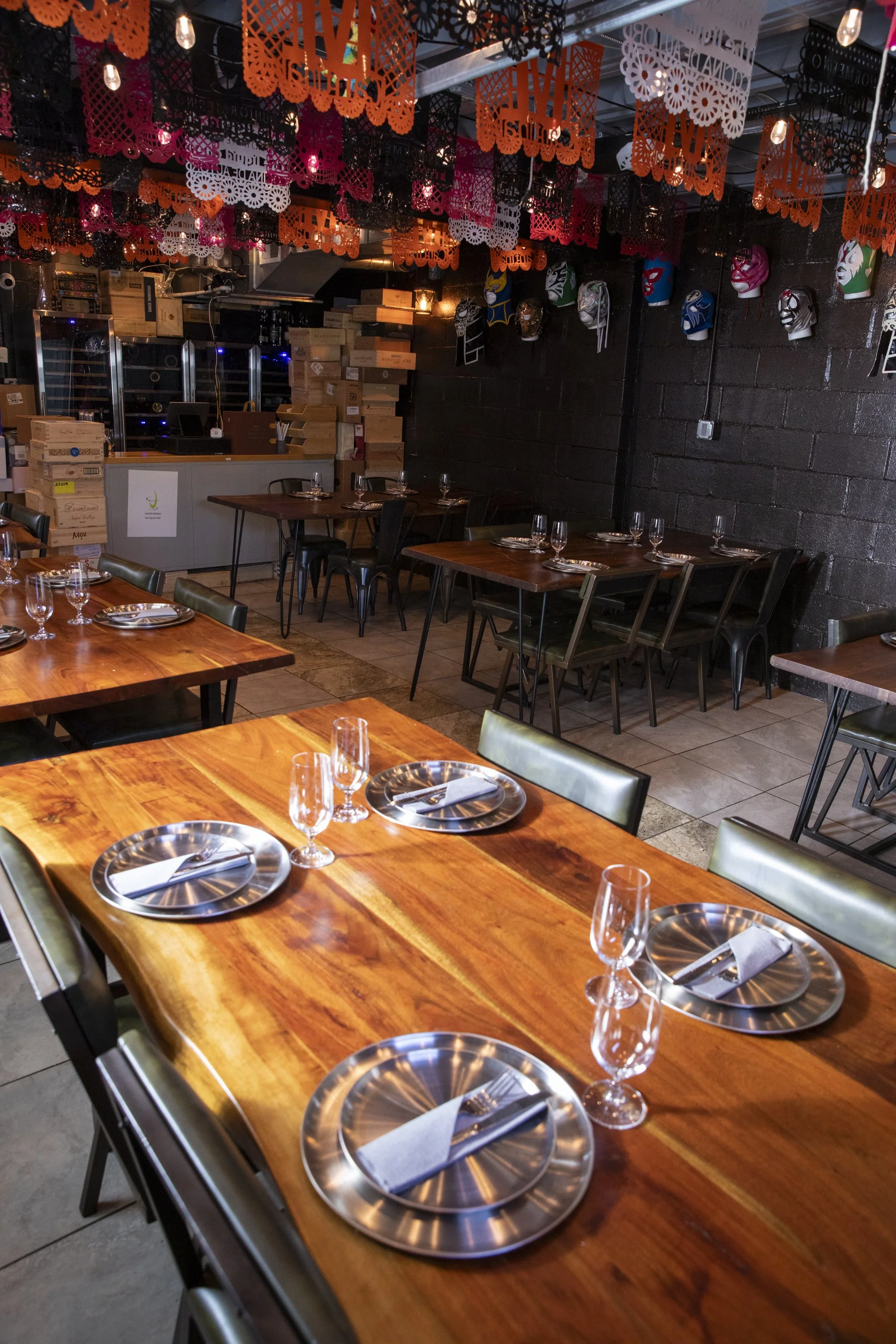 A restaurant interior decorated with colorful papel picado banners hanging from the ceiling and luchador masks on the black brick wall, with wooden tables set with glassware and napkins.