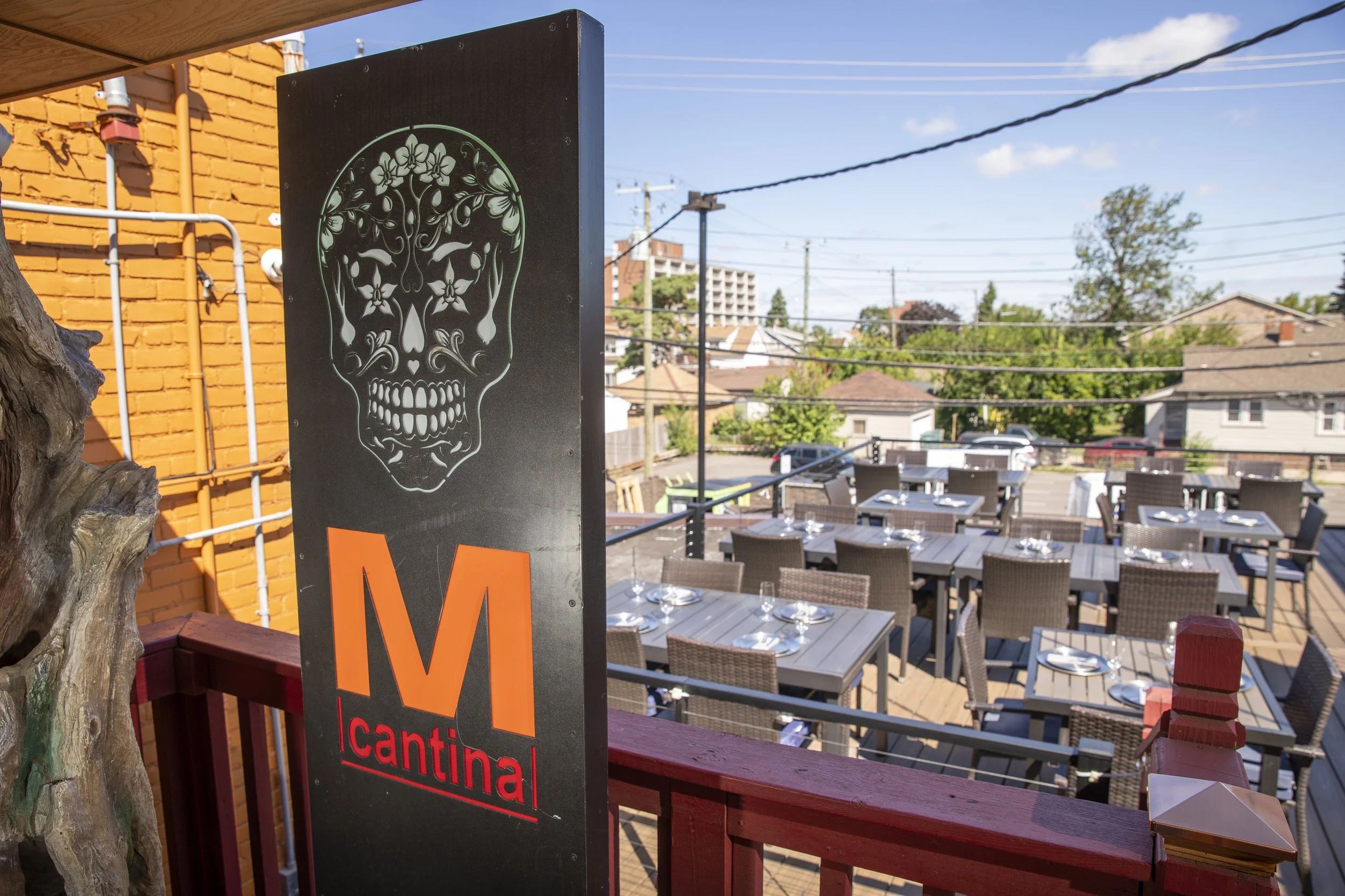 Outdoor dining area with tables set with glasses, covered by a pergola. A black sign with a decorative skull and the word 'cantina' in red letters is visible, with a brick wall and utility poles in the background.