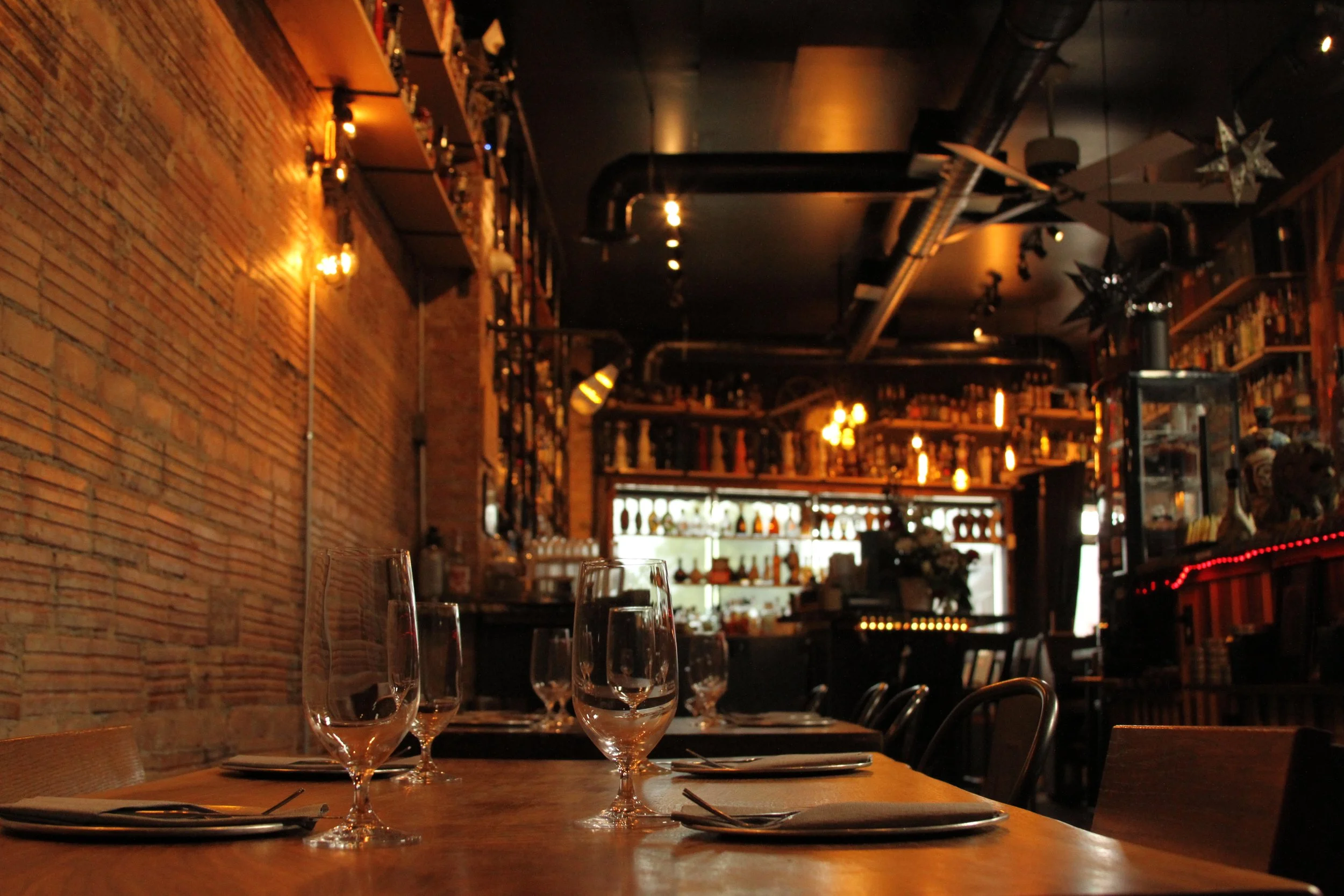 Interior of a dimly lit restaurant with brick wall, empty wine glasses on table, and shelves with bottles and decorative stars hanging from the ceiling.