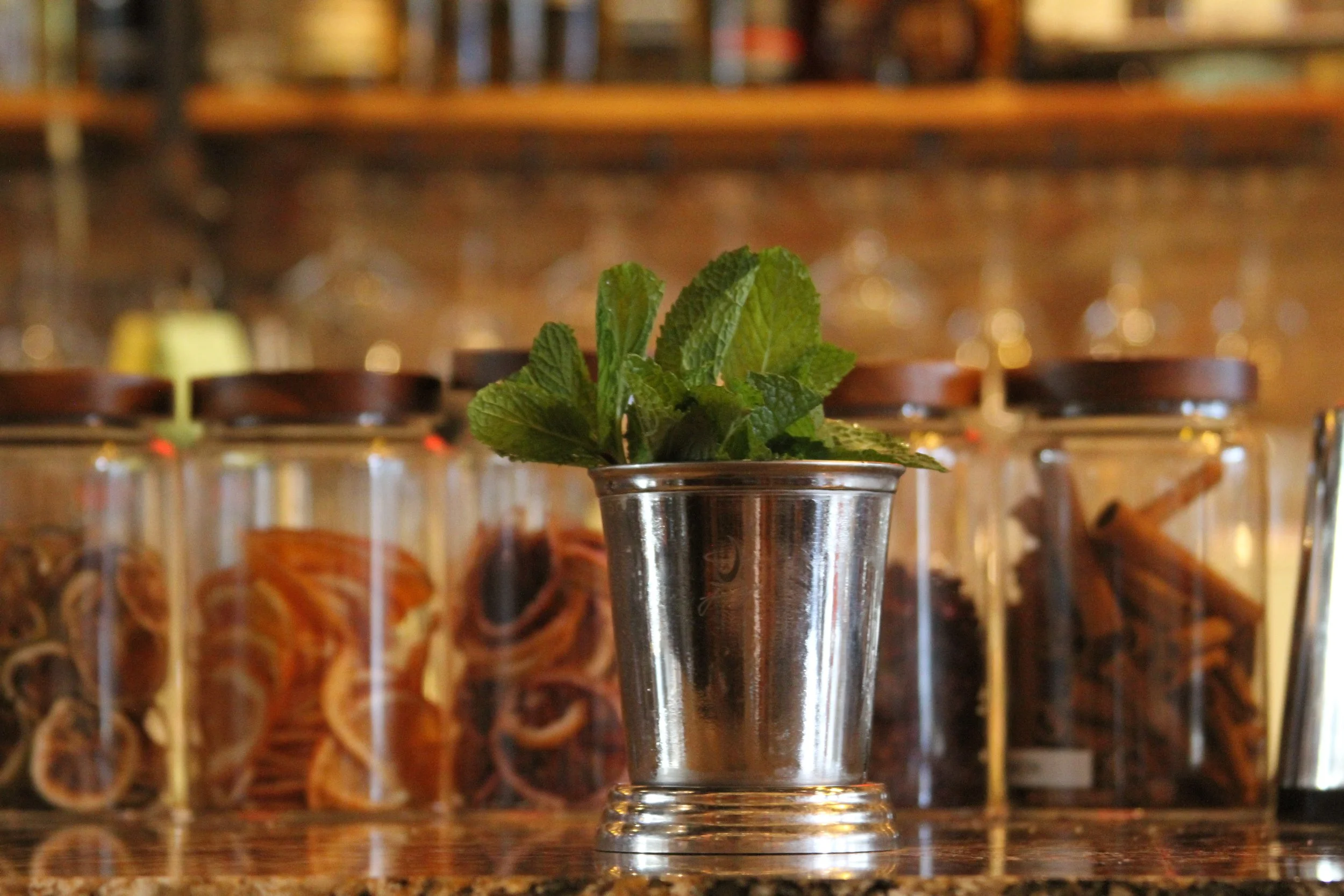 Fresh mint leaves in a metal cocktail shaker on a counter with jars of dried herbs and spices in the background.