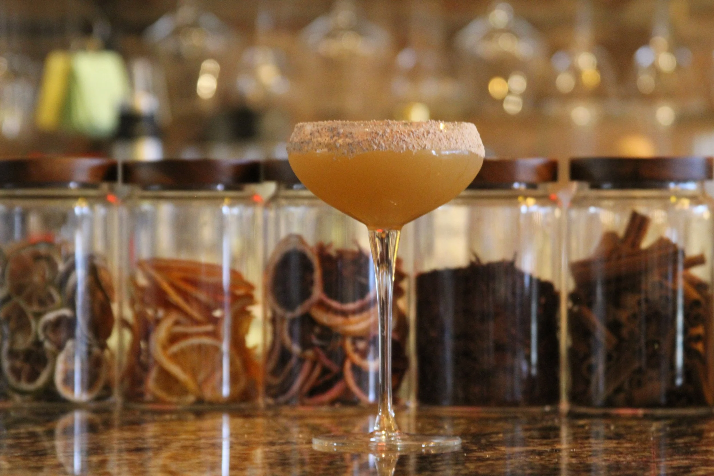 A cocktail with a salted rim in a wine glass, set on a bar counter with blurred bottles and decor in the background.