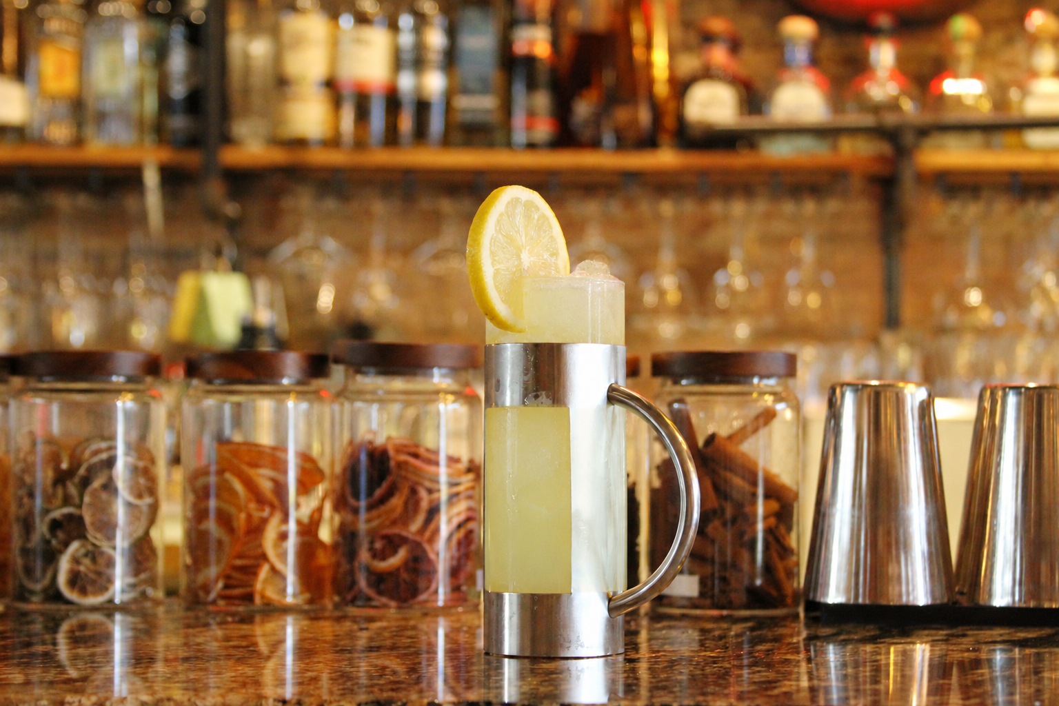 A cocktail with a lemon slice garnish on top, placed on a bar counter with jars of dried citrus slices and spices in the background.