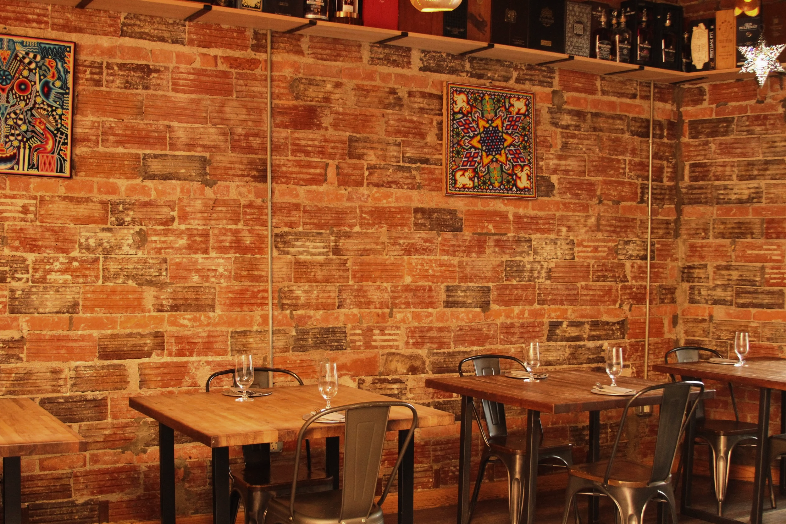 A cozy restaurant interior with exposed brick walls decorated with colorful, vibrant artwork. Wooden tables are set with wine glasses and napkins, and black metal chairs surround the tables. Shelves with bottles and decorative star ornaments are mounted on the wall.