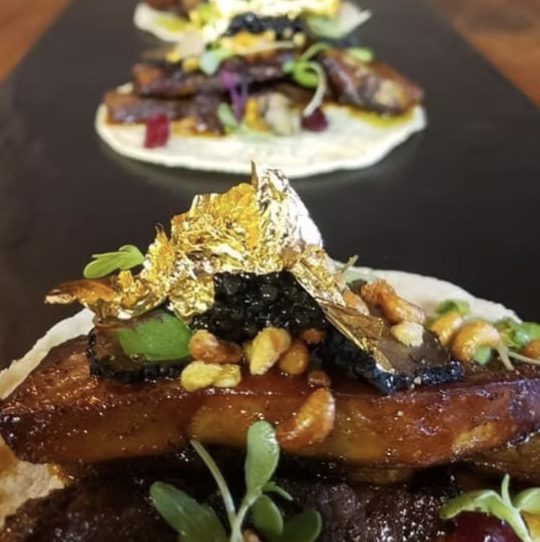Close-up of a gourmet open-faced sandwich with glazed meat, topped with edible gold leaf, black caviar, nuts, herbs, and vegetables on a white bread slice.