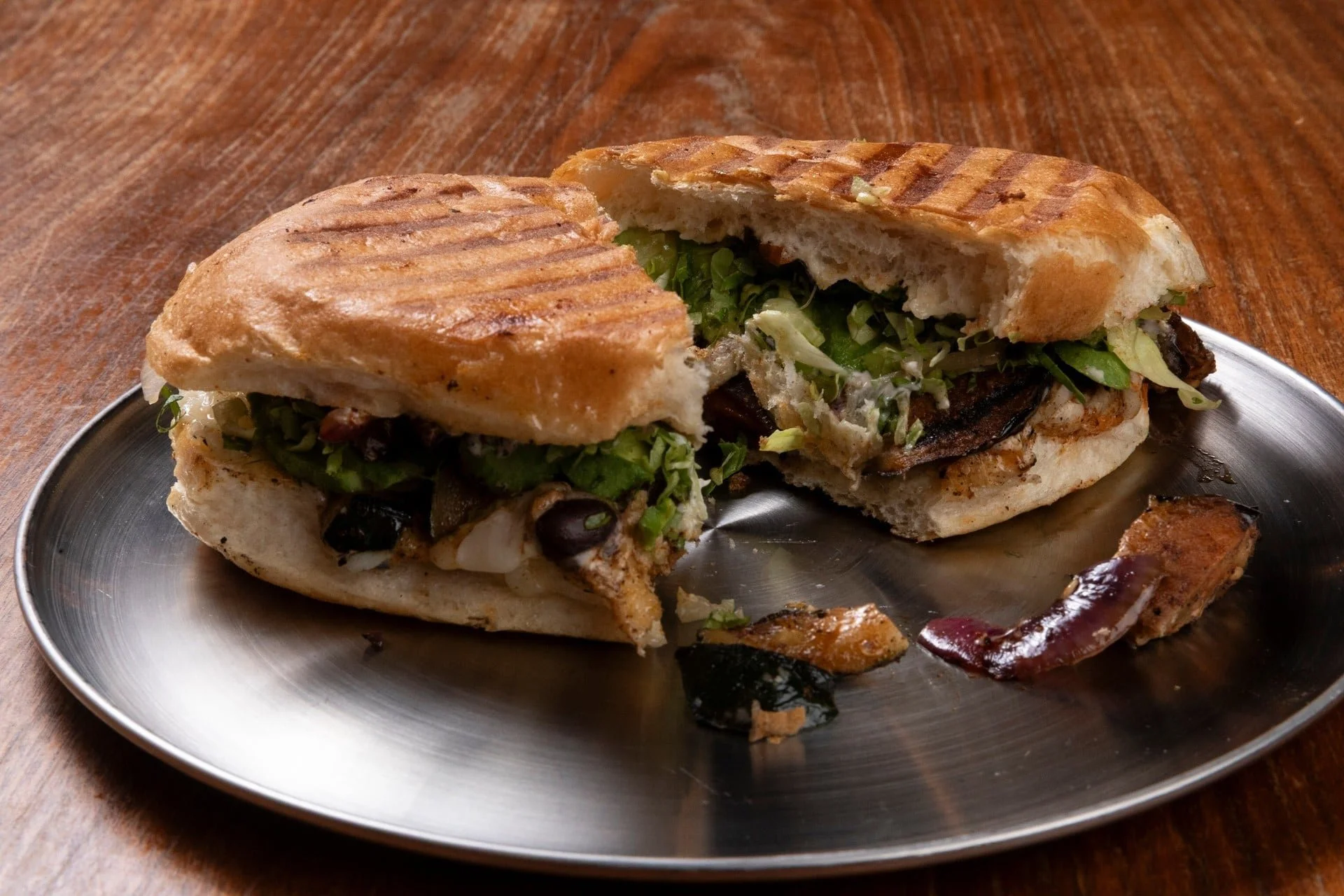 Close-up of a grilled sandwich with lettuce, black beans, and melted cheese on a metal plate, with a side of grilled meat and a wooden table in the background.