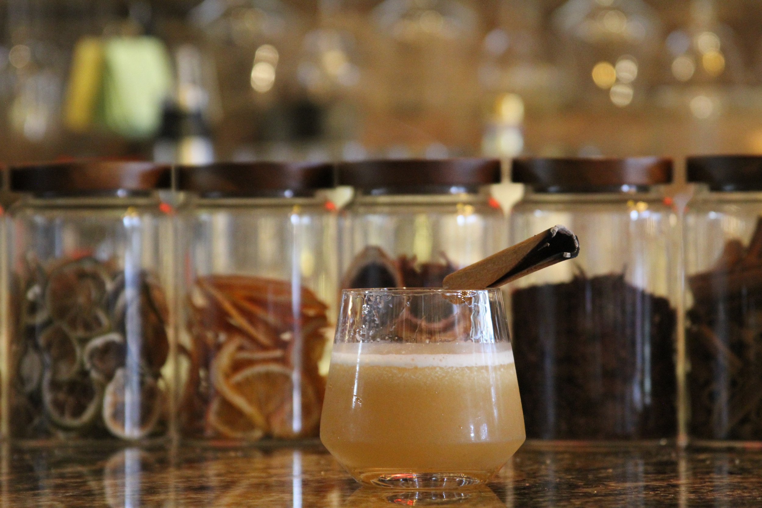 A glass of beer with a frothy head on a counter in front of a blurred background of spice jars and seasonings.