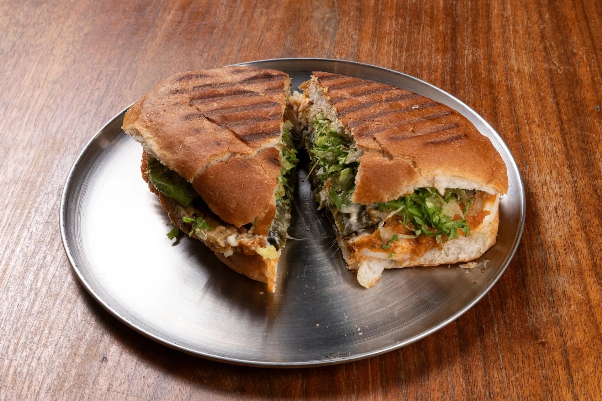 Half-eaten hoagie sandwich on a metal plate, with lettuce, vegetables, and sauce, on a wooden table.