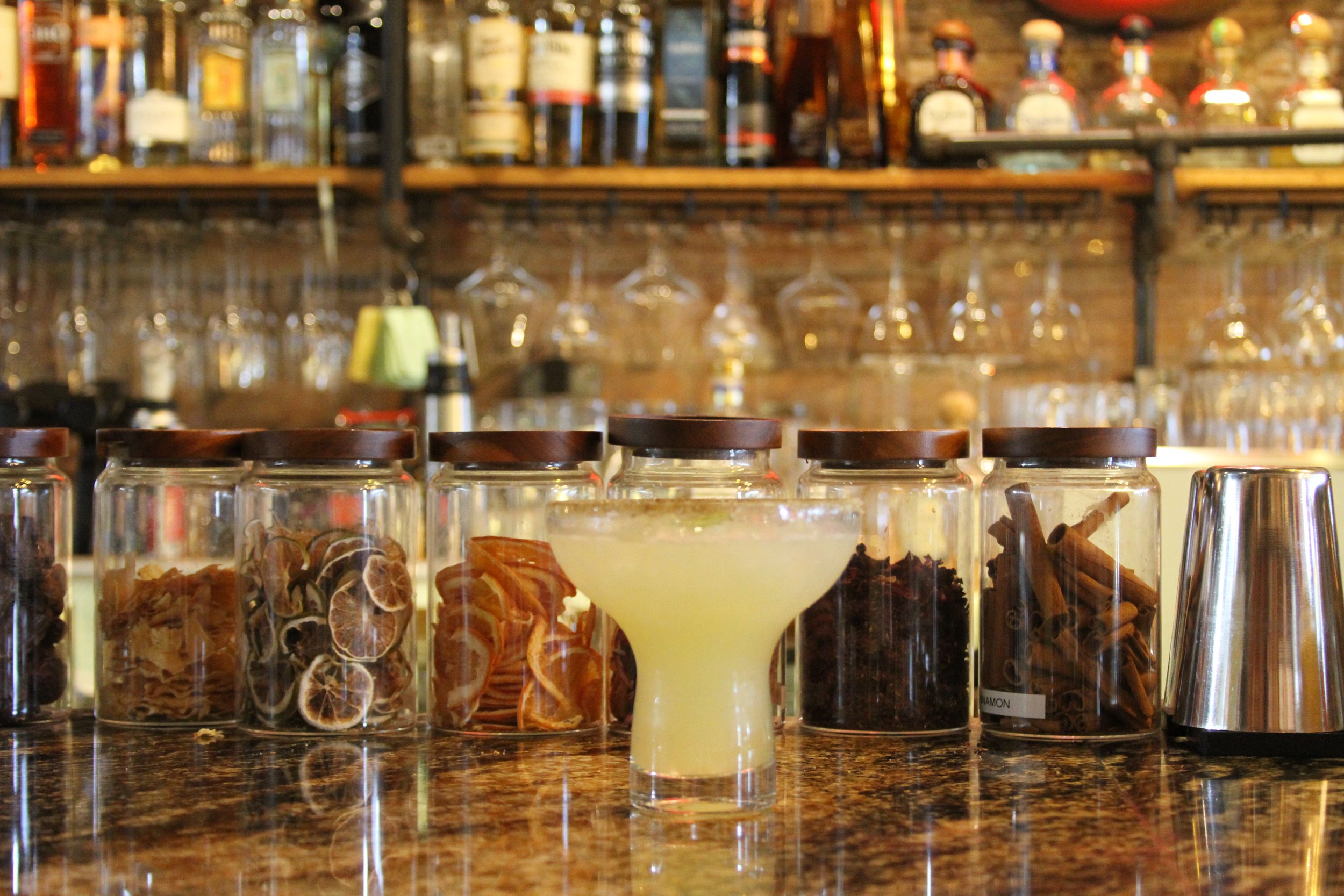 A row of glass jars with wooden lids containing dried herbs and spices, placed on a dark countertop in front of a bar with various bottles and hanging glasses.