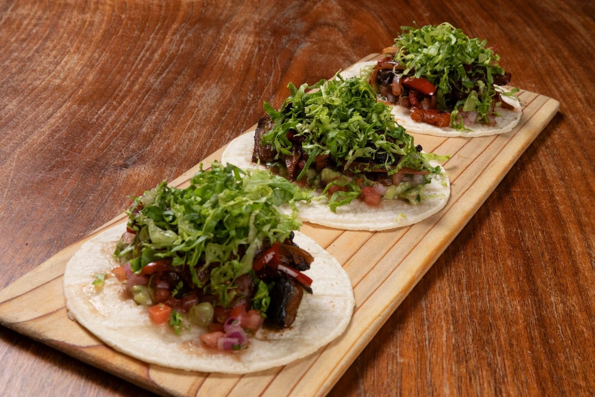 Three tacos with chopped meat, lettuce, and other vegetables on a wooden board.