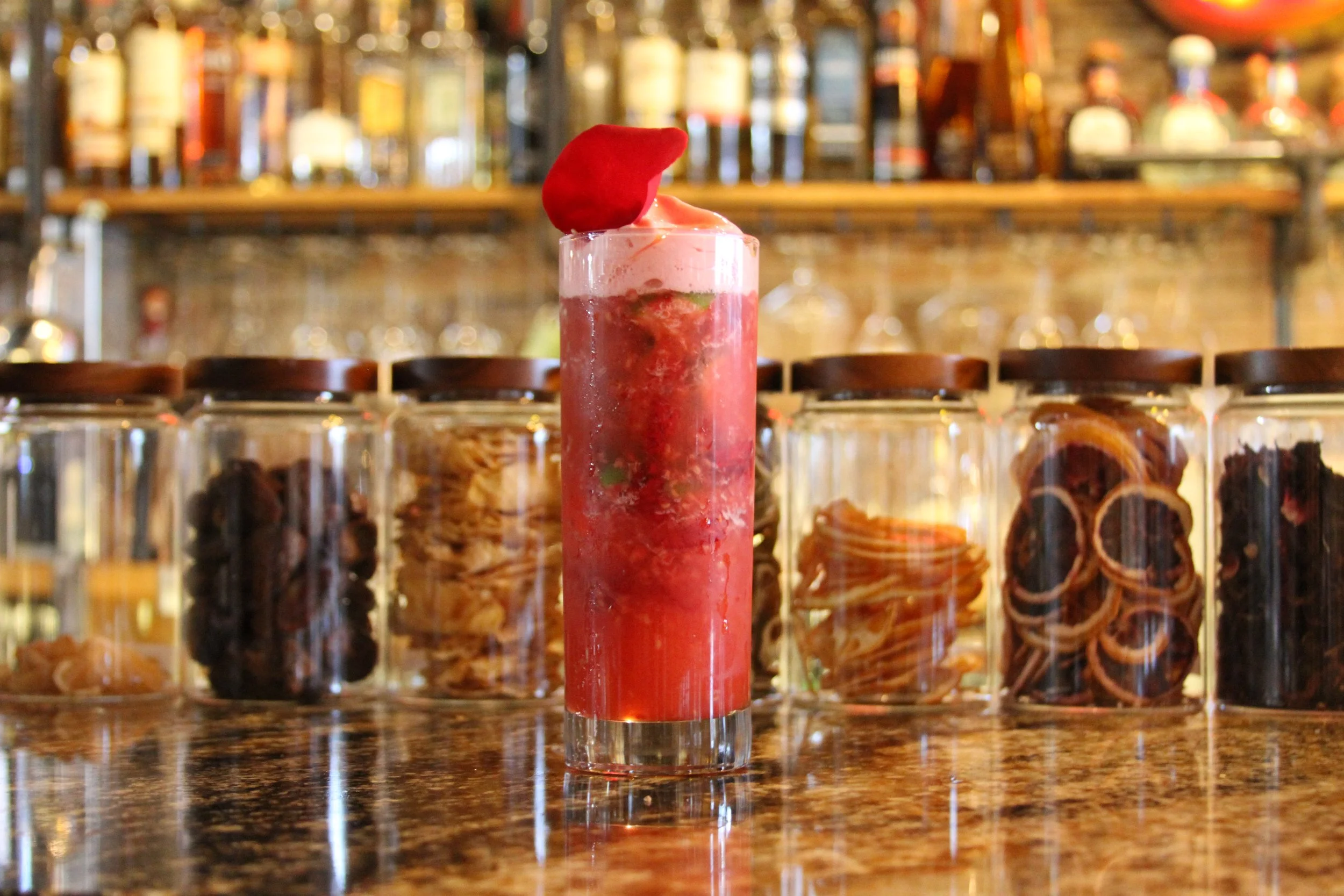 A tall, clear glass filled with a layered red and pink cocktail, garnished with a red flower petal, placed on a dark, polished countertop with a blurred bar in the background.